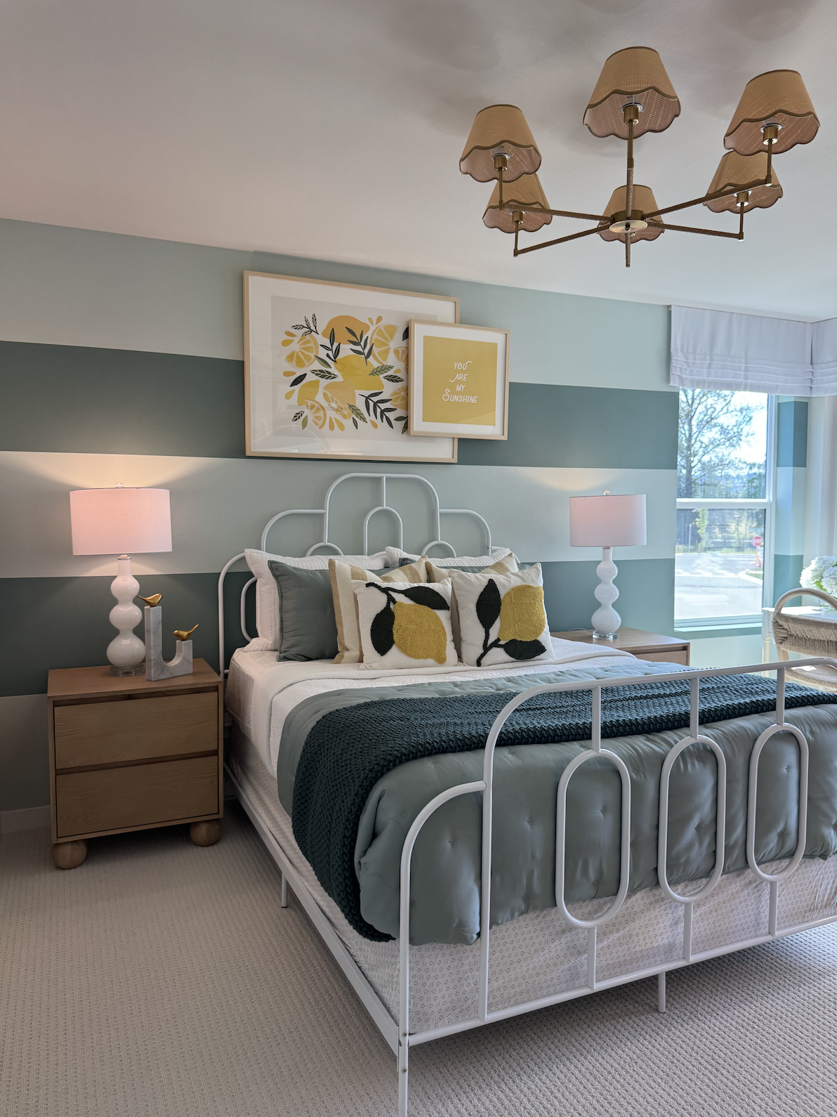 A cozy bedroom with a white metal bed frame, featuring yellow, black, and white decorative pillows with lemon and leaf motifs. The room has two nightstands with white lamps, three framed art pieces above the bed, one with an abstract lemon design and another with the phrase 'You are my sunshine.' There are two windows, a beige carpet, and a mid-century modern chandelier with six beige shades hanging from the ceiling.