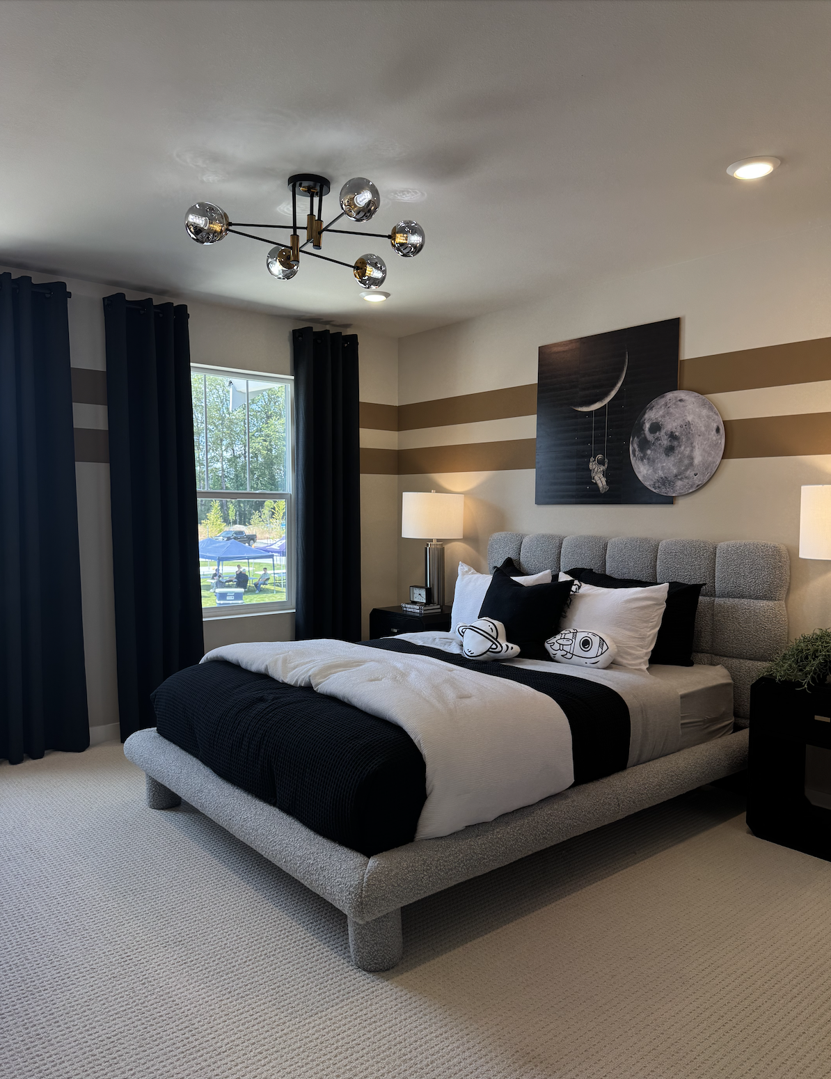 Modern bedroom with a large bed, space-themed artwork on the wall, black and white bedding with space-themed pillows, dark curtains, and a window showing a backyard with outdoor chairs and umbrellas.