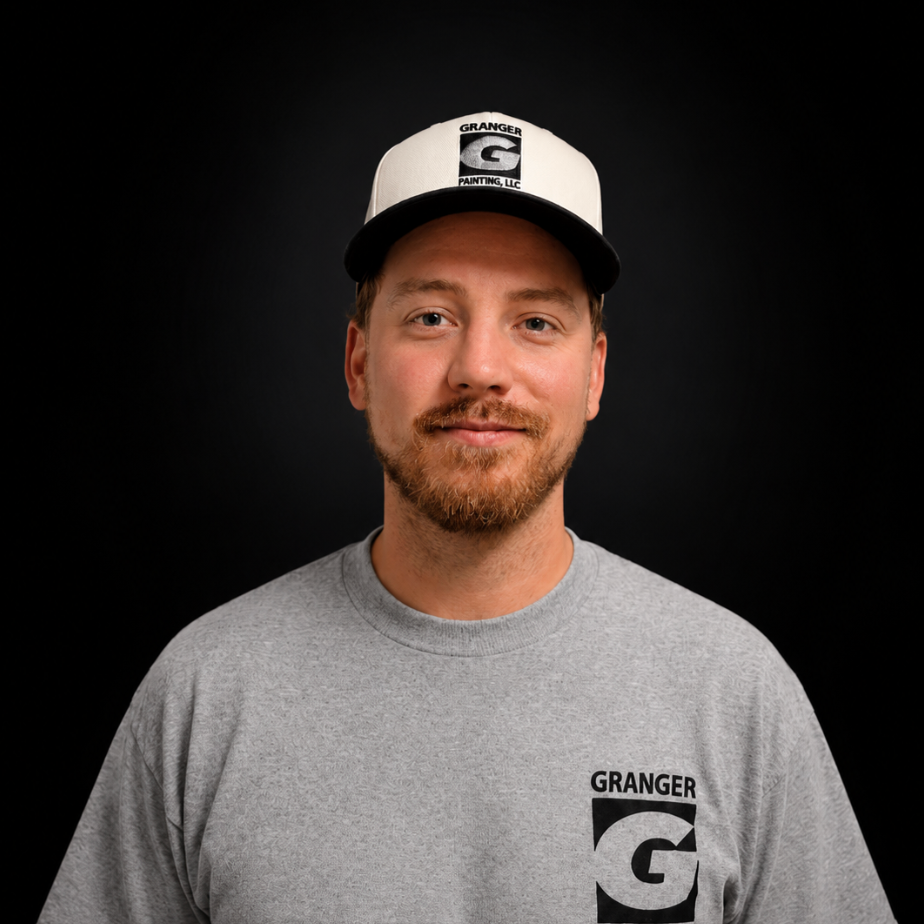 A man with light skin, red hair, and a beard wearing a white and black cap and a gray t-shirt with the logo 'GRANGER G Painting, LLC' on both the cap and shirt, standing against a black background.