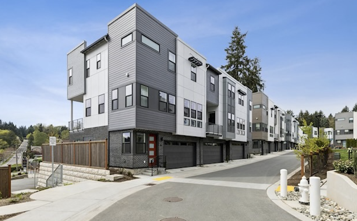 Modern multi-story residential buildings with garages at street level, situated on a gently curved road, with landscaped areas and a partly cloudy sky in the background.