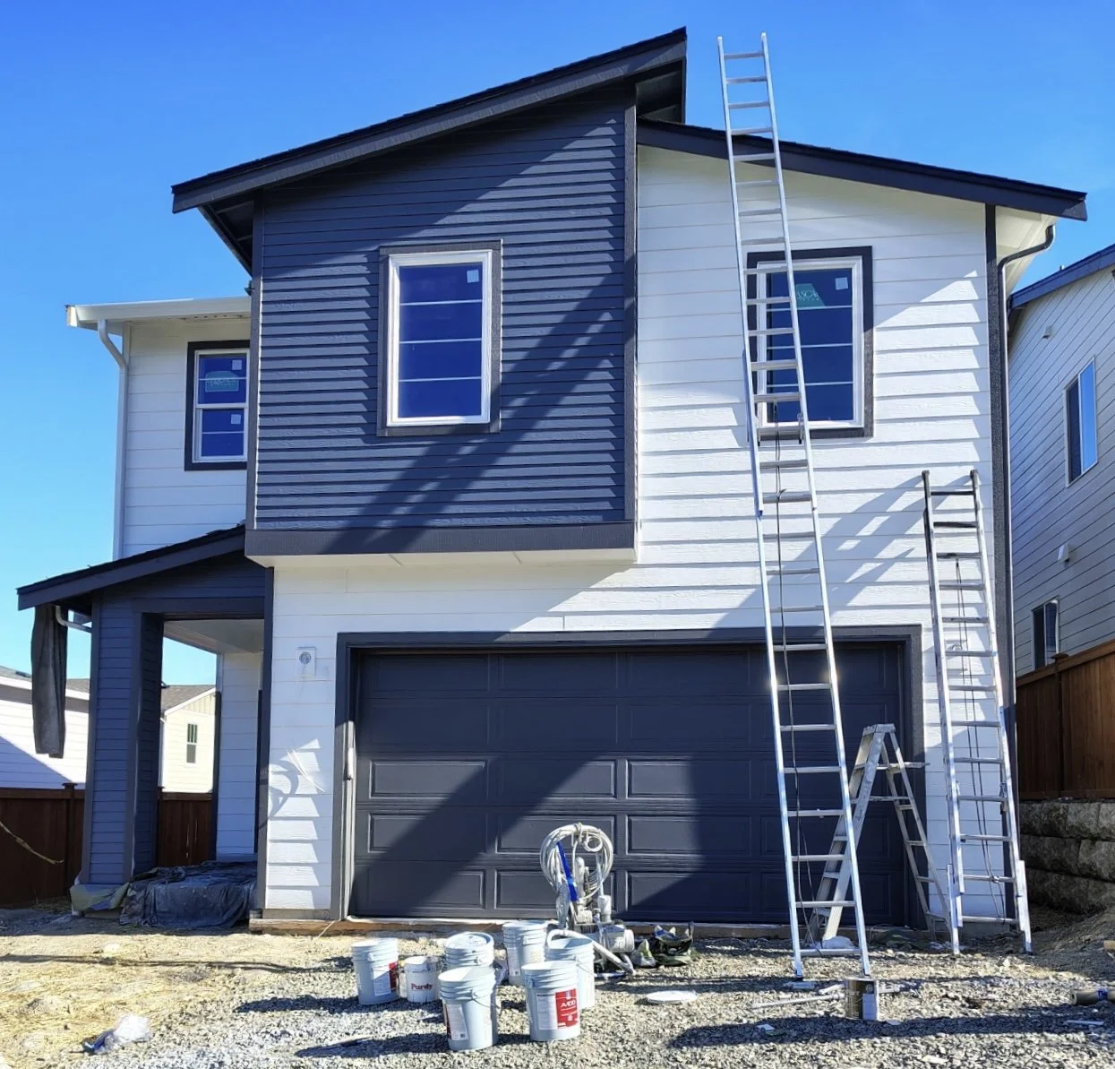 Two-story house under construction that is being painted, ladders leaning against the house, and buckets of paint or construction materials on the ground.