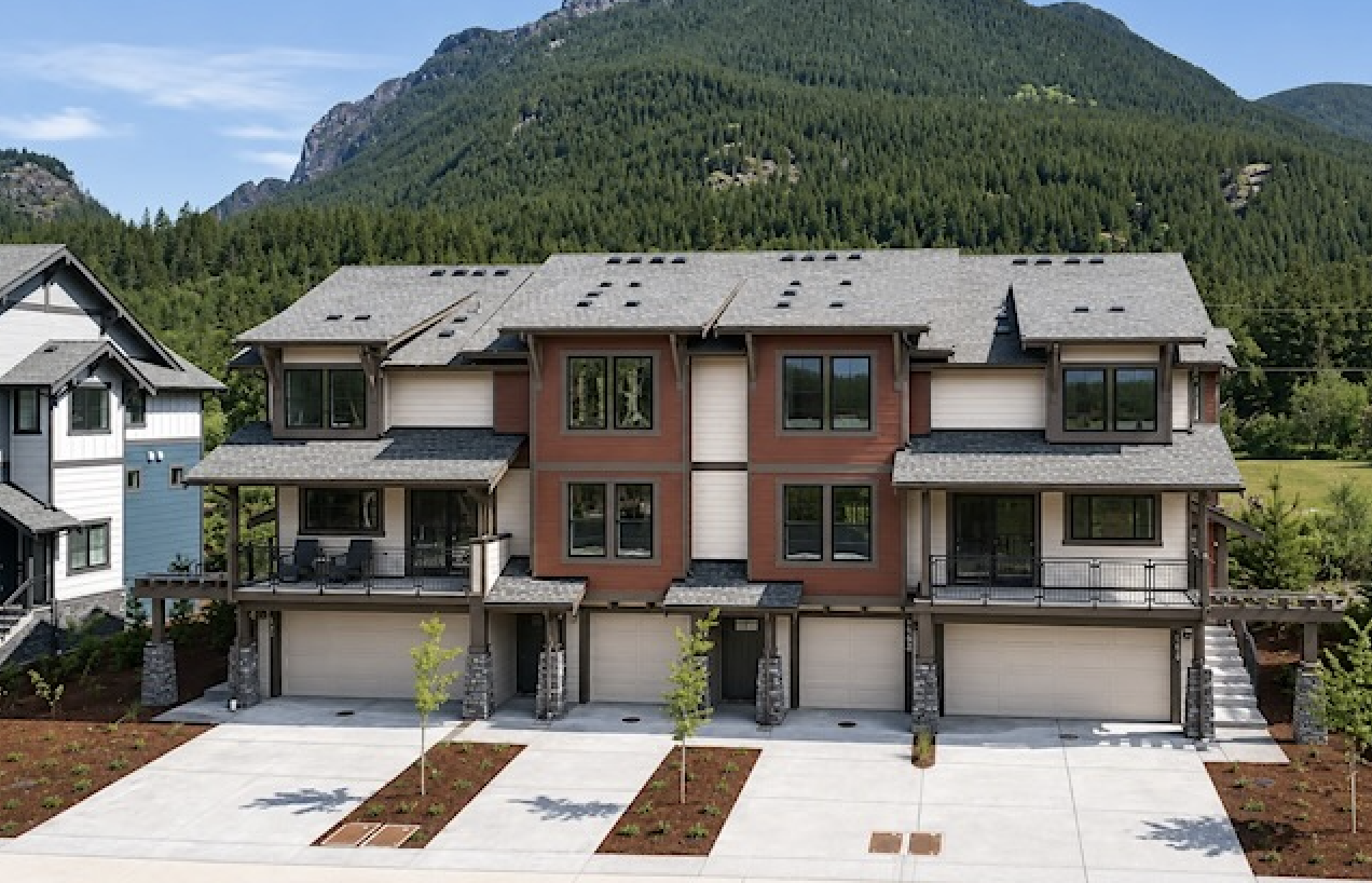 A modern three-story multifamily with a garage, balconies, and large windows, set against a mountainous and forested background.