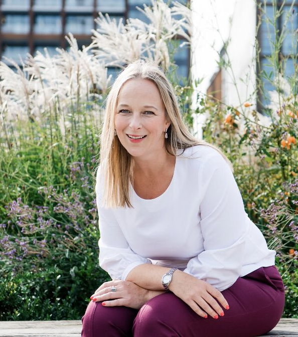 A woman with blonde hair sitting on a bench in a garden with various flowering plants, wearing a white blouse and maroon pants.