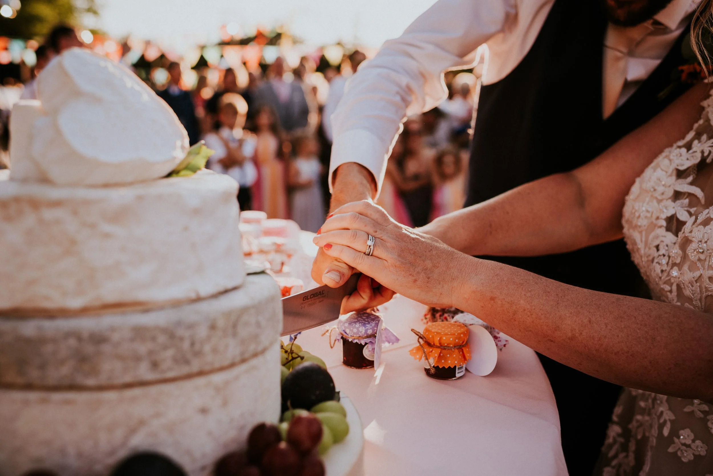 Close-up of a person cutting a cake at an outdoor celebration, with people and colorful decorations in the background.