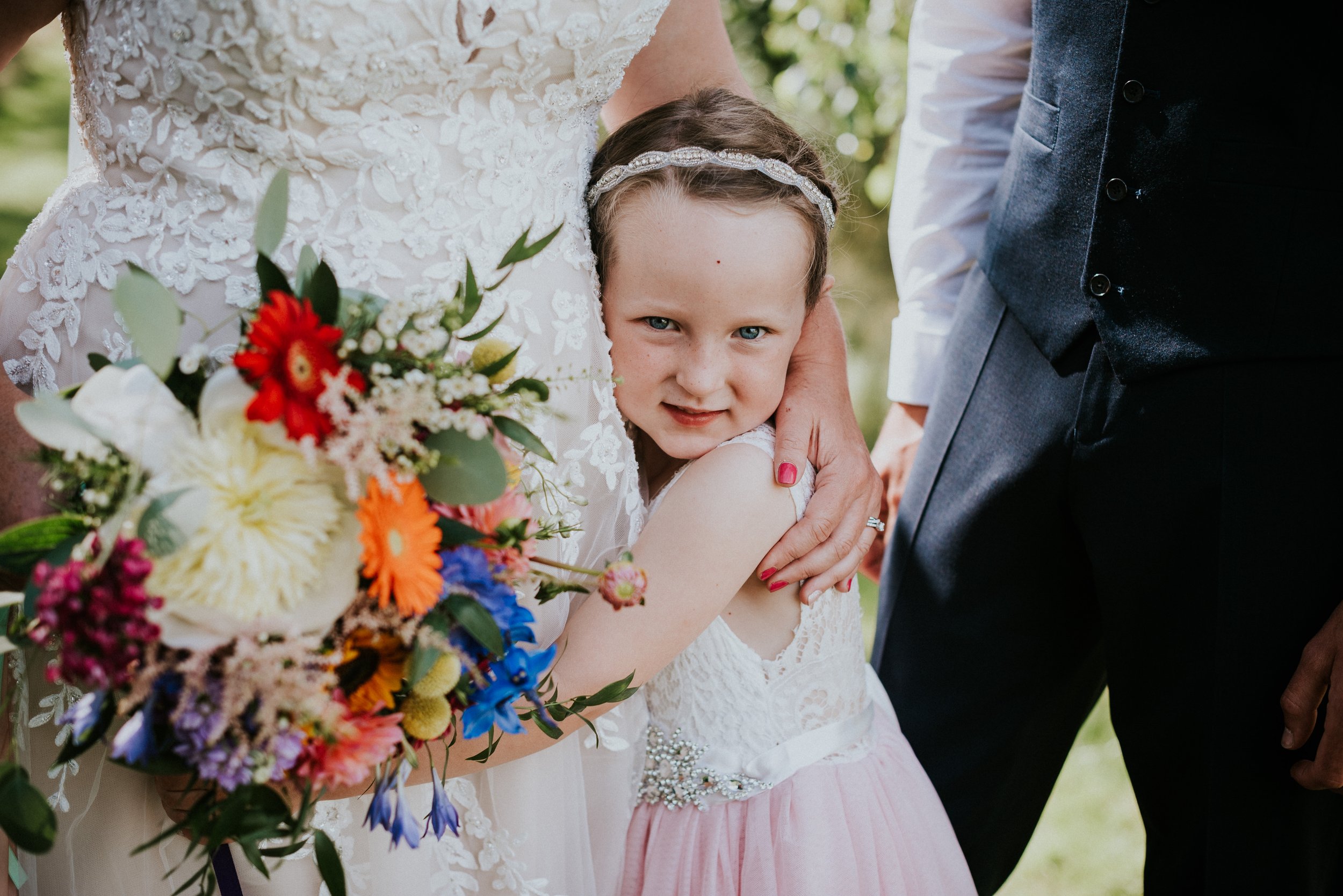 A young girl with blue eyes and light skin, wearing a white dress and a silver headband, is being hugged by a woman in a white lace dress holding a colorful bouquet of flowers, with a man in a dark vest and white shirt standing nearby outdoors.