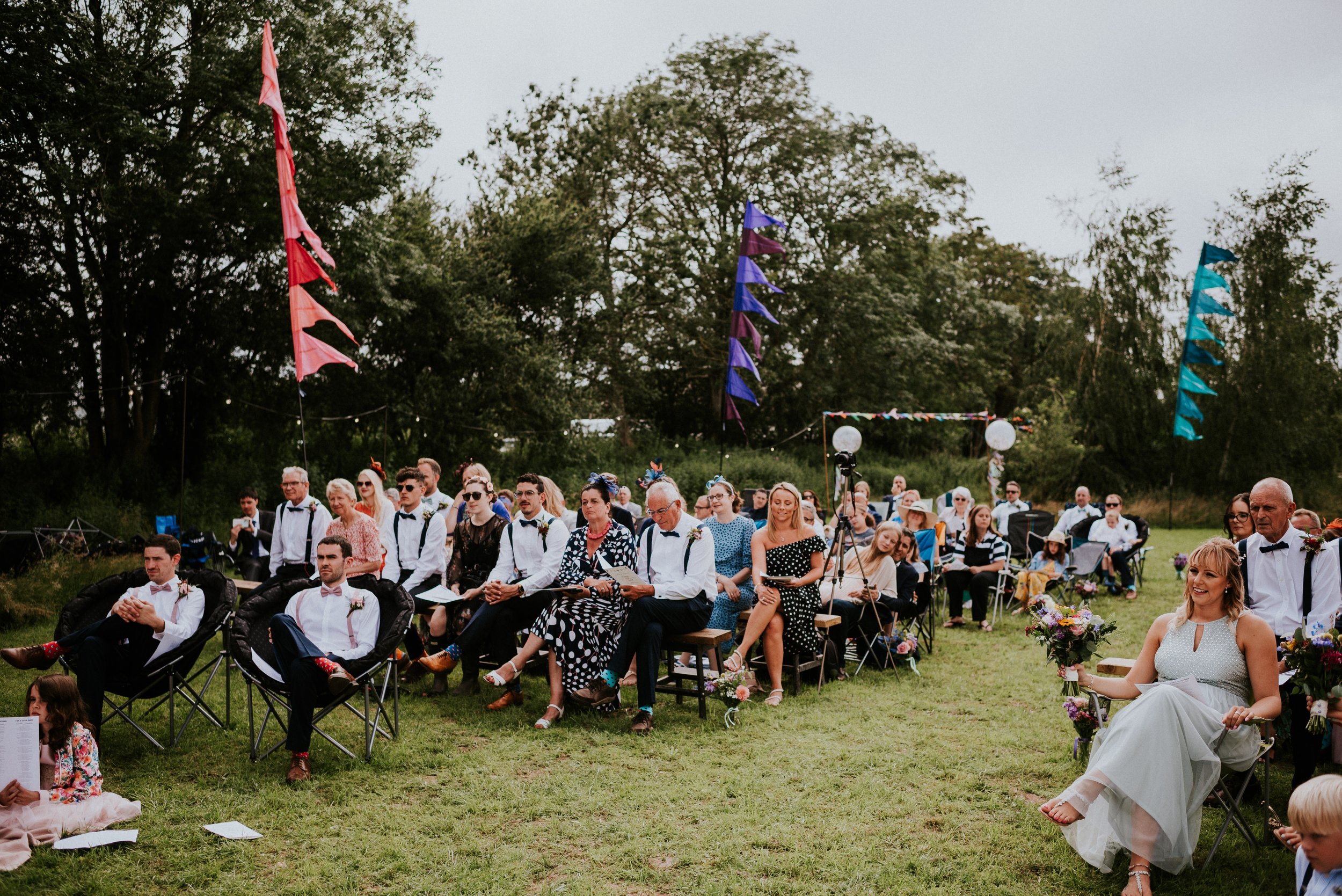 Outdoor wedding ceremony with guests seated on chairs, colorful flags and trees in the background.