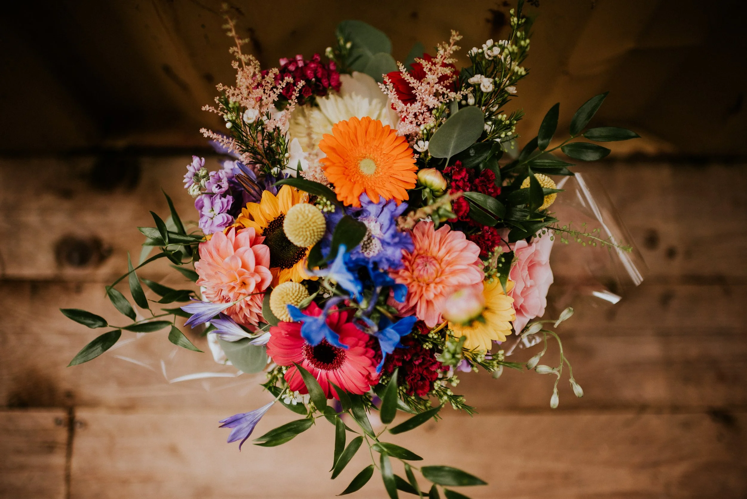 Colorful flower bouquet with orange, pink, purple, white, yellow, and red flowers alongside green leaves on a wooden surface.
