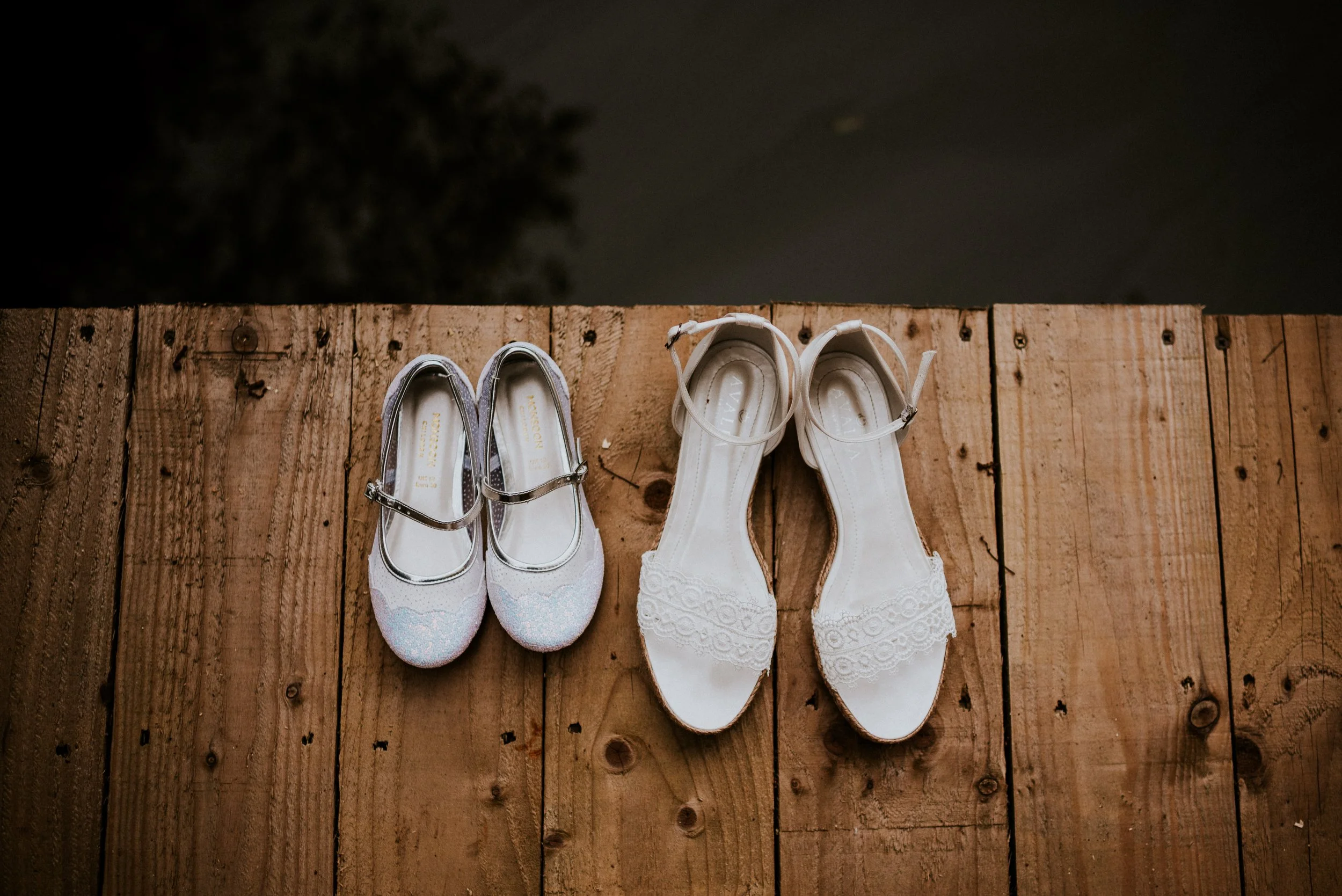 Two pairs of shoes, one with sparkly flats and the other with white lace-up heels, placed on a wooden surface outdoors at night.