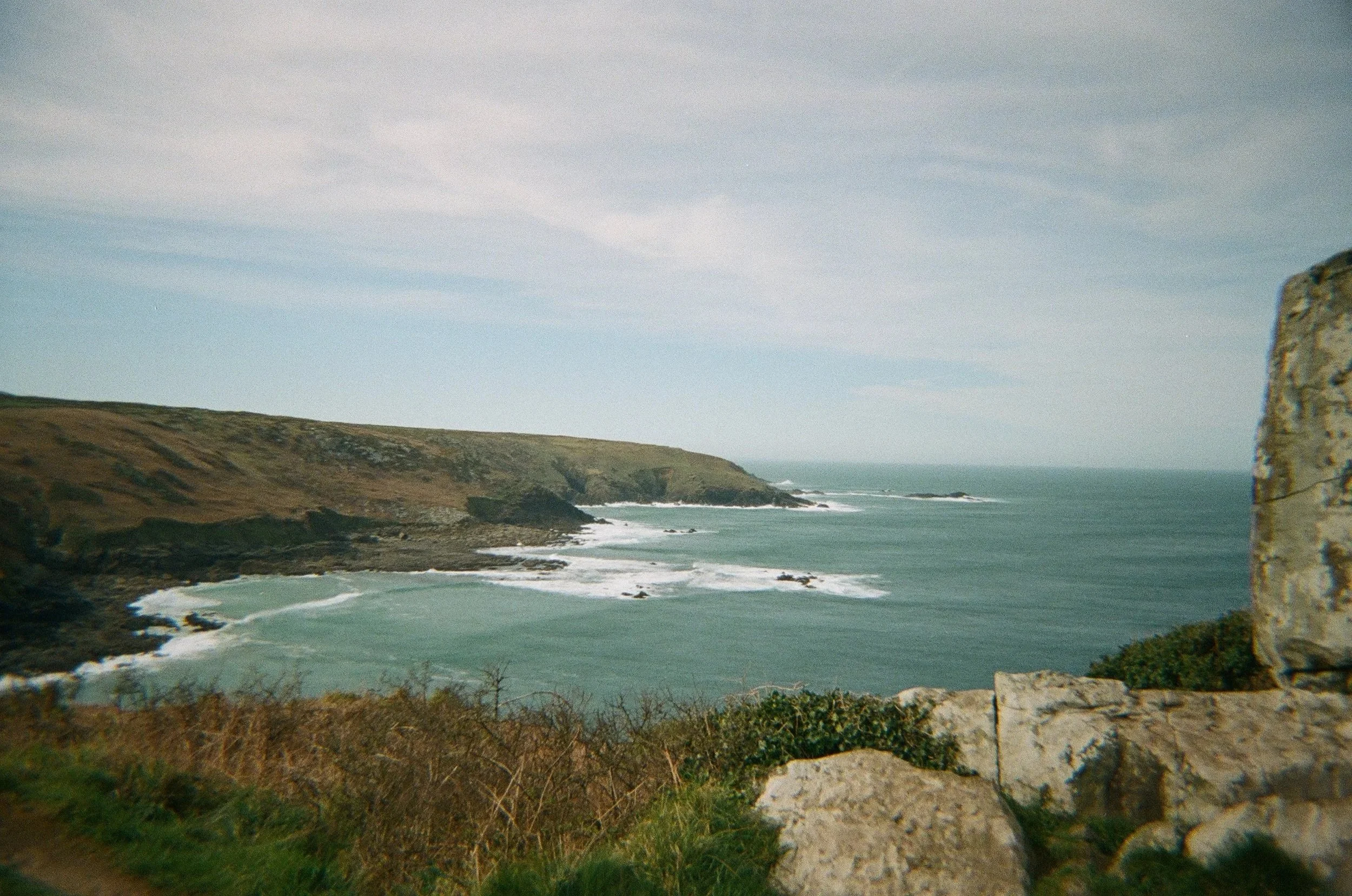 Scenic view of a coastline with cliffs, ocean waves, and a rocky outcrop on the right, with cloudy sky above.