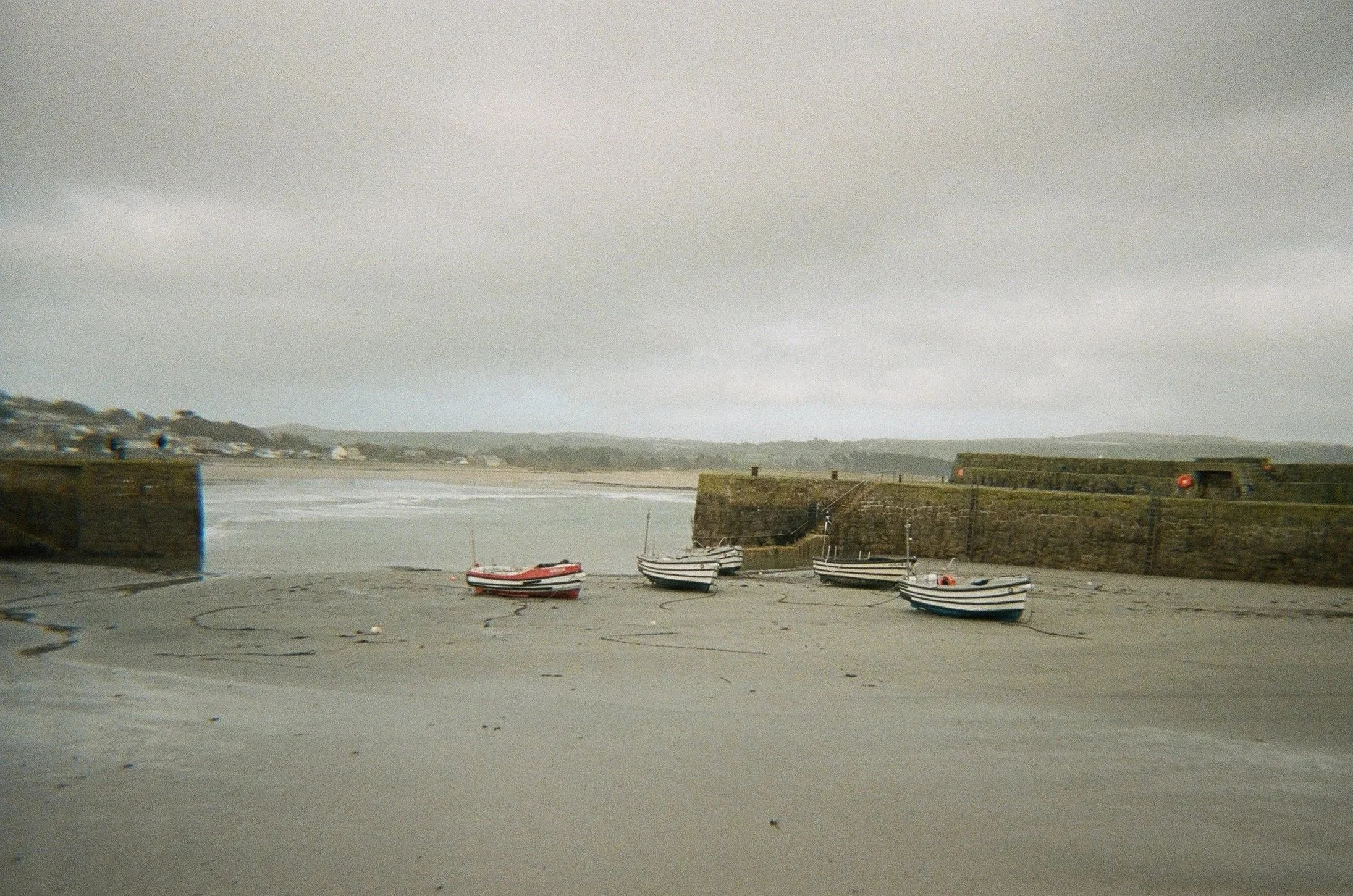 A harbor scene with four small boats resting on the concrete ground near a stone pier, with a body of water and a distant hill in the background under a cloudy sky.