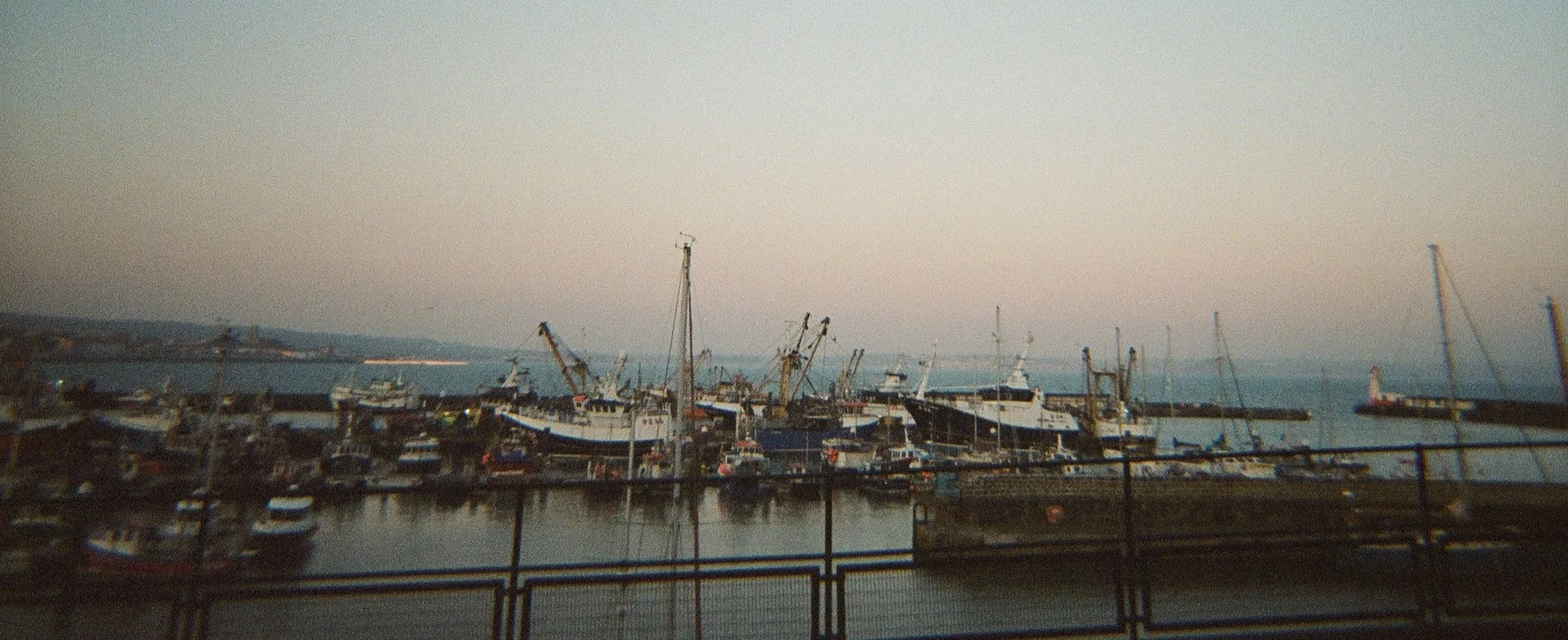 A harbor with multiple boats and ships docked, some with masts, during dusk or dawn, with calm water and a distant shoreline.