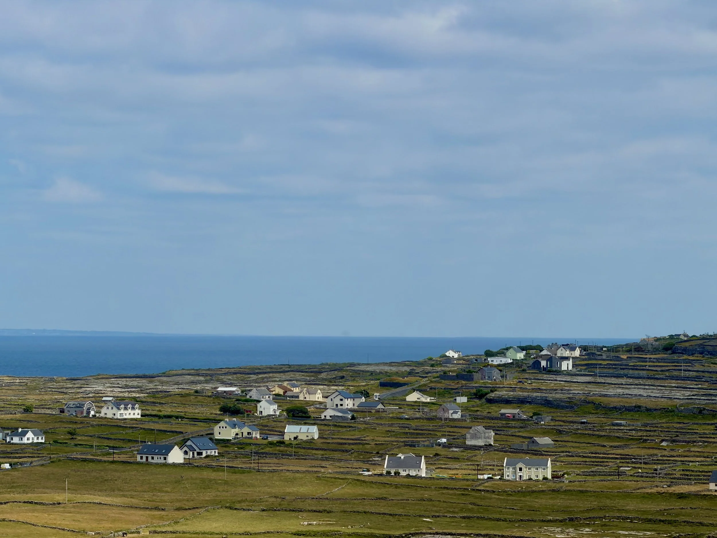 A seaside landscape with a body of water and a partly cloudy sky. There are several houses and buildings scattered across the land near the coast.