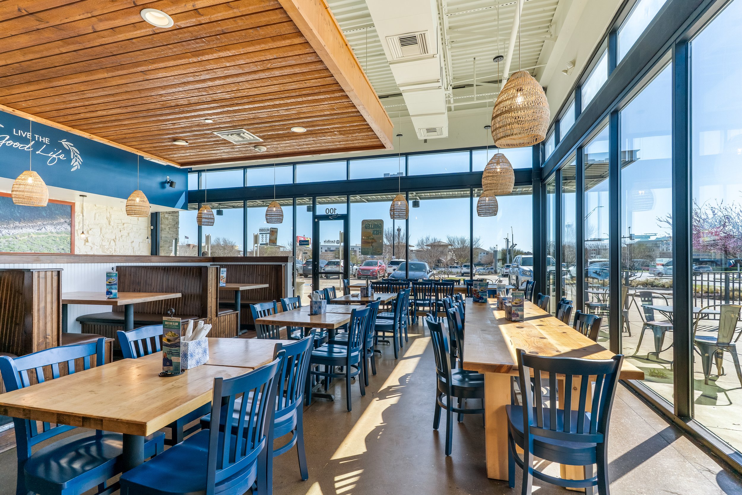 Interior of a modern restaurant with wooden ceiling, blue walls, and large windows showing parking lot outside. The dining area features blue chairs and wooden tables, with hanging wicker lamps.