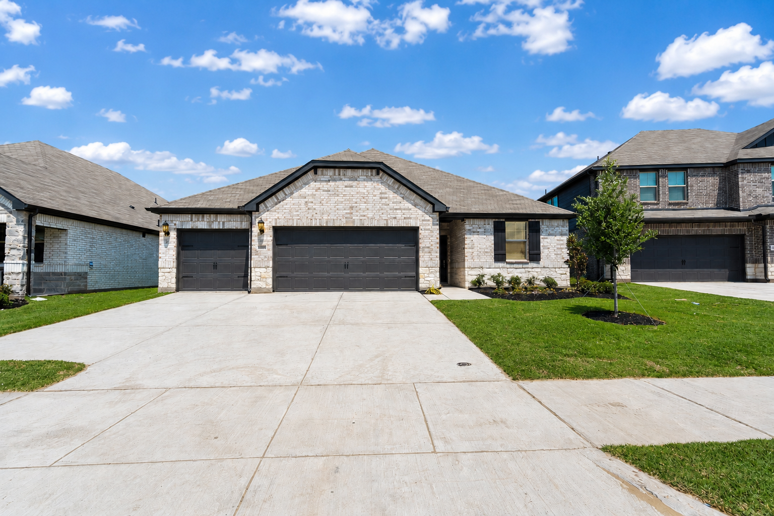 Modern house with three garages, brick exterior, black garage doors, and small front yard with green lawn and young tree under a blue sky with scattered clouds.