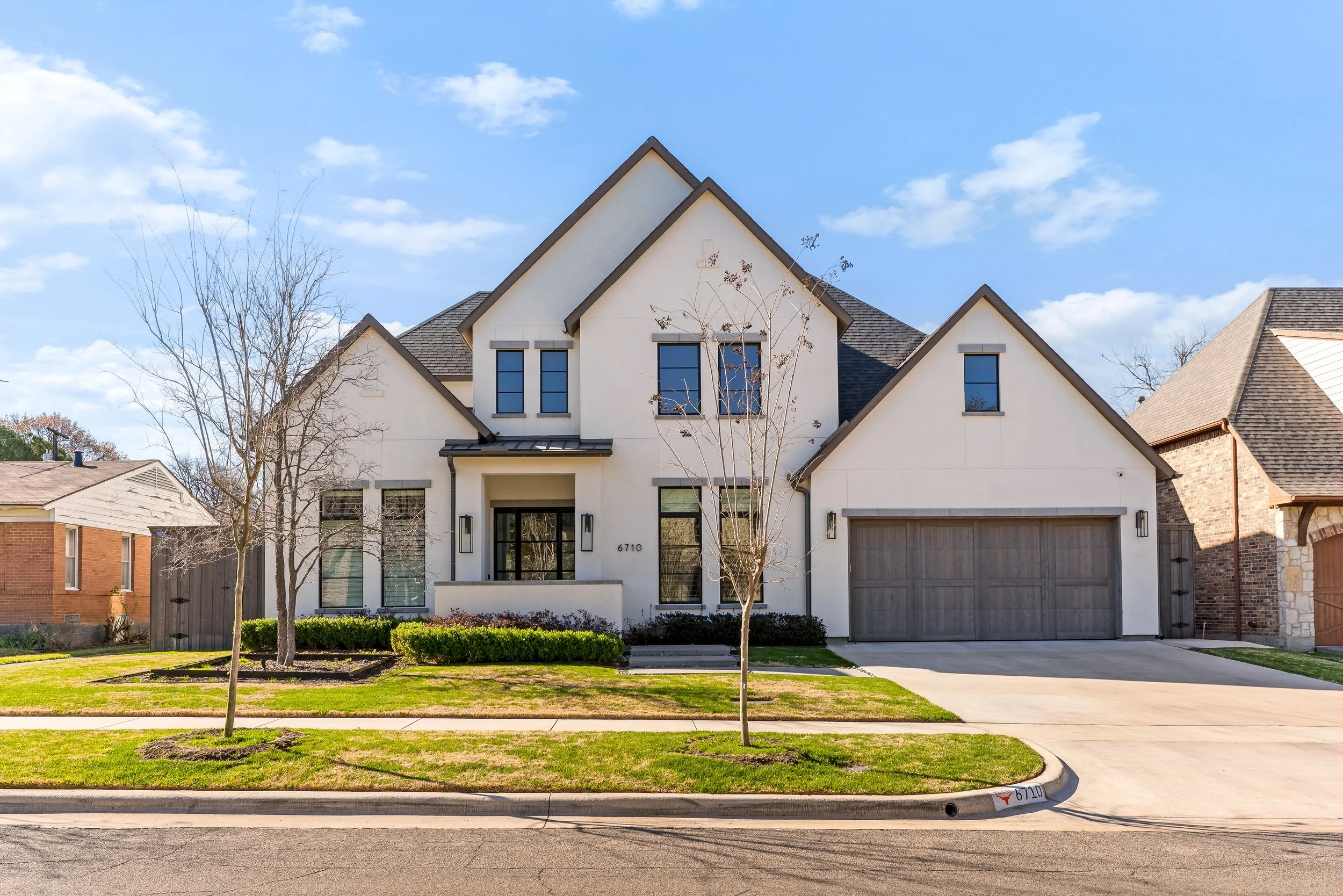Front view of a modern two-story house with a white exterior, black window frames, and a gray roof. There are two leafless trees in the front yard, a manicured lawn, and a concrete driveway leading to a garage. The sky is clear with scattered clouds.