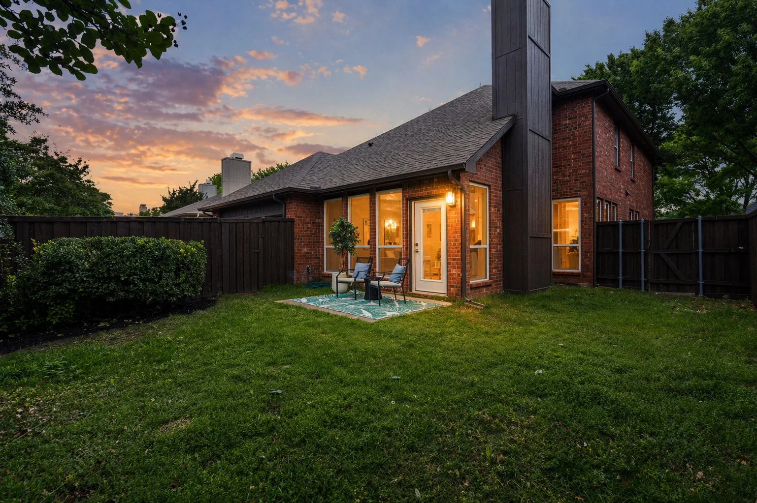 Backyard of a brick house at sunset with a small patio area, two chairs, a small table, a patterned rug, and a fence surrounding the yard.