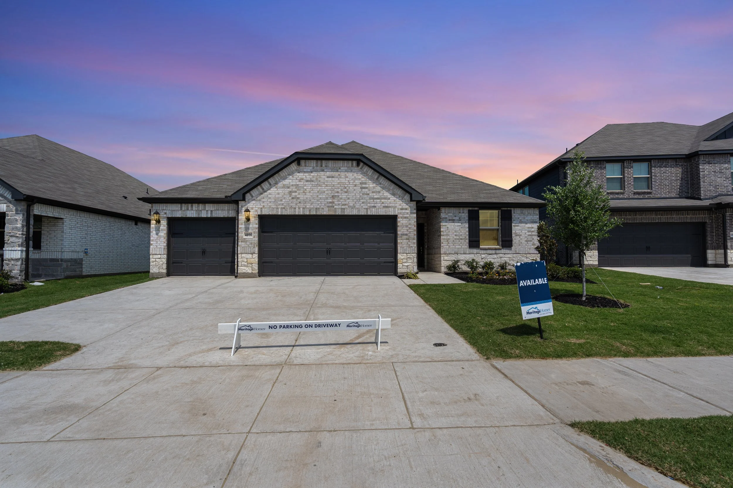 Image of new house with brick exterior and new lawn, with a sign that indicates 'No parking on driveway' and an 'Available' sign from Meritage Homes, at sunset with purple and pink sky.