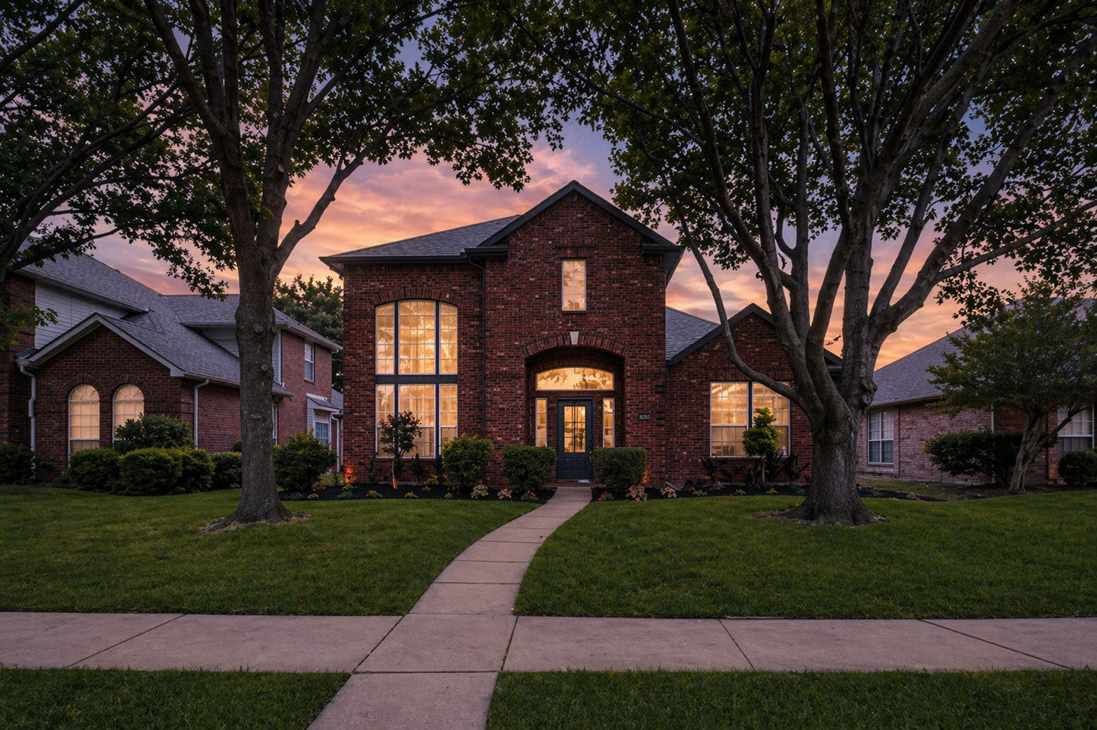 A large brick house with tall windows glowing with warm light during sunset, surrounded by a green lawn and trees, with a front walkway leading to the door.
