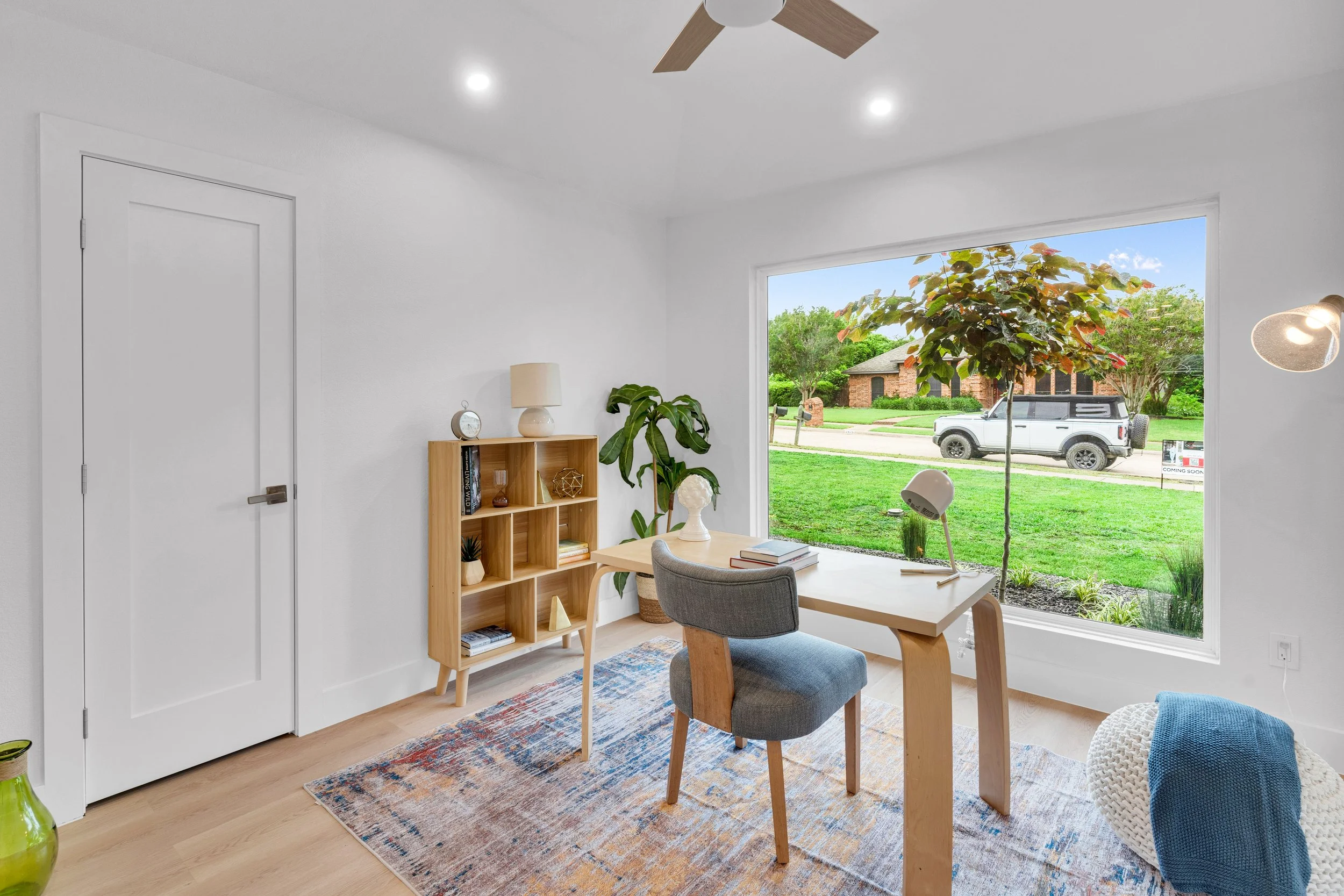 A room with a large window overlooking a green lawn and trees. Inside, there's a wooden dining table with a chair, a bookshelf, a potted plant, a table lamp, and a decorative bust. The floor has a colorful rug, and a ceiling fan is visible on the cei