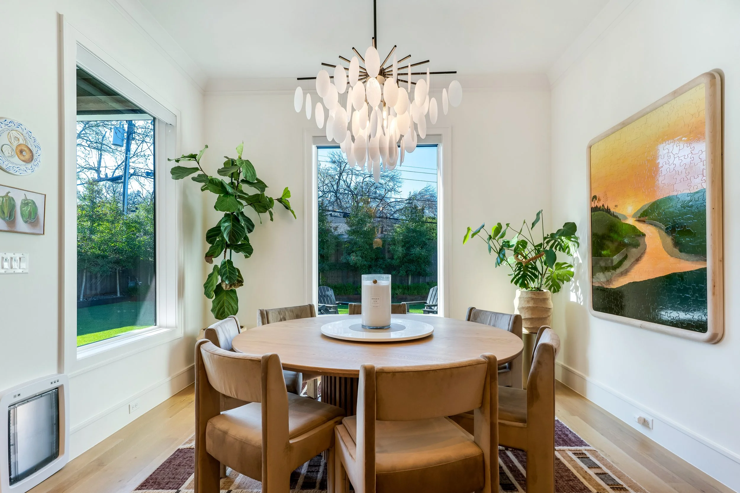 Dining room with a round wooden table surrounded by tan chairs, large windows showing trees outside, a white chandelier hanging from the ceiling, indoor plants, and a framed landscape painting on the wall.