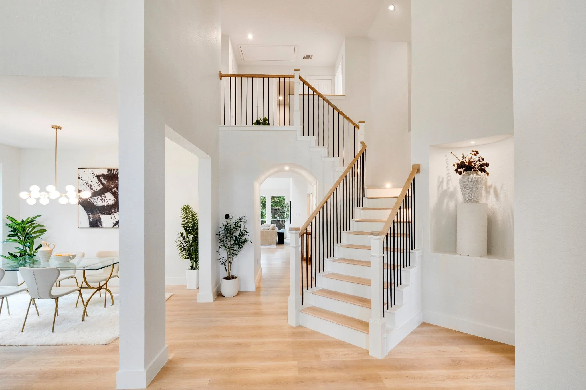 Interior view of a modern, well-lit home with a staircase featuring black railings and wooden steps. The space has white walls, hardwood floors, and decor including large plants and artwork. A dining area with white chairs and a glass table is visibl