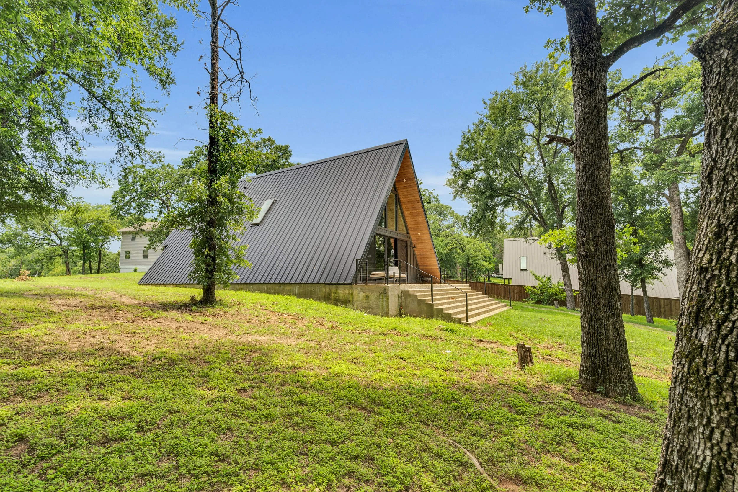 A modern A-frame house with dark gray metal roof and wooden accents, situated in a lush green area with large trees and grass, with some steps leading up to a small porch.