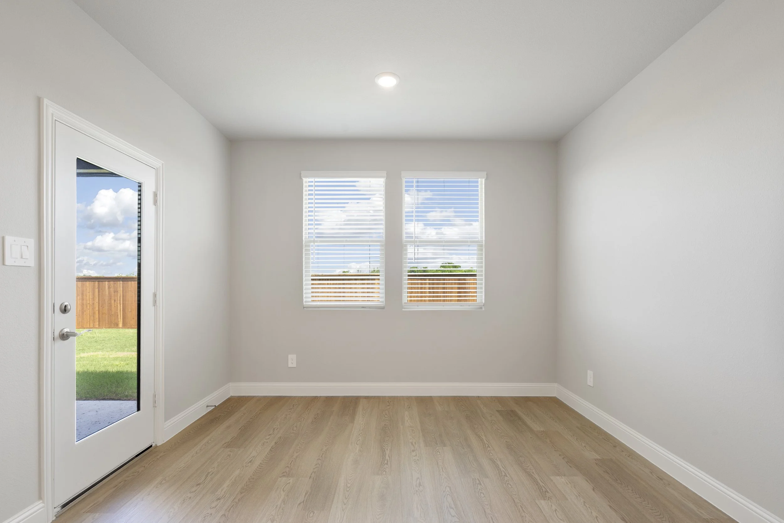 Empty room with white walls, hardwood floor, two windows with blinds, a door leading outside, and ceiling light.