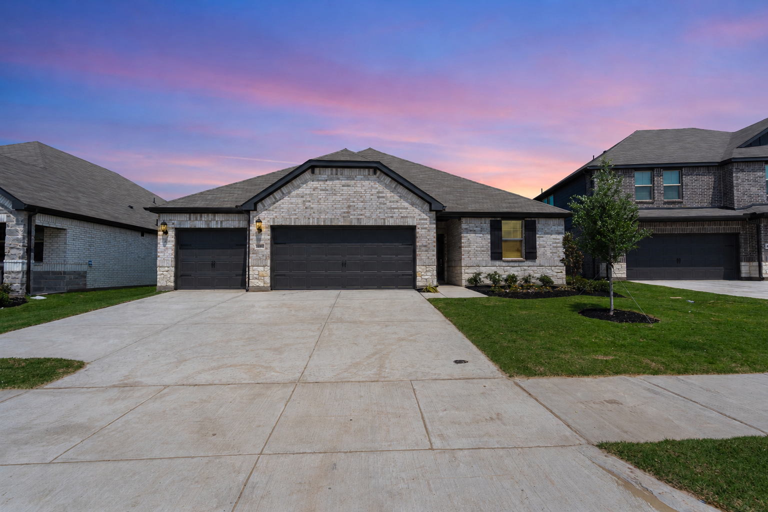 Front view of a modern brick house with a two-car garage, front yard with grass and a small tree, and a colorful sunset sky in the background.