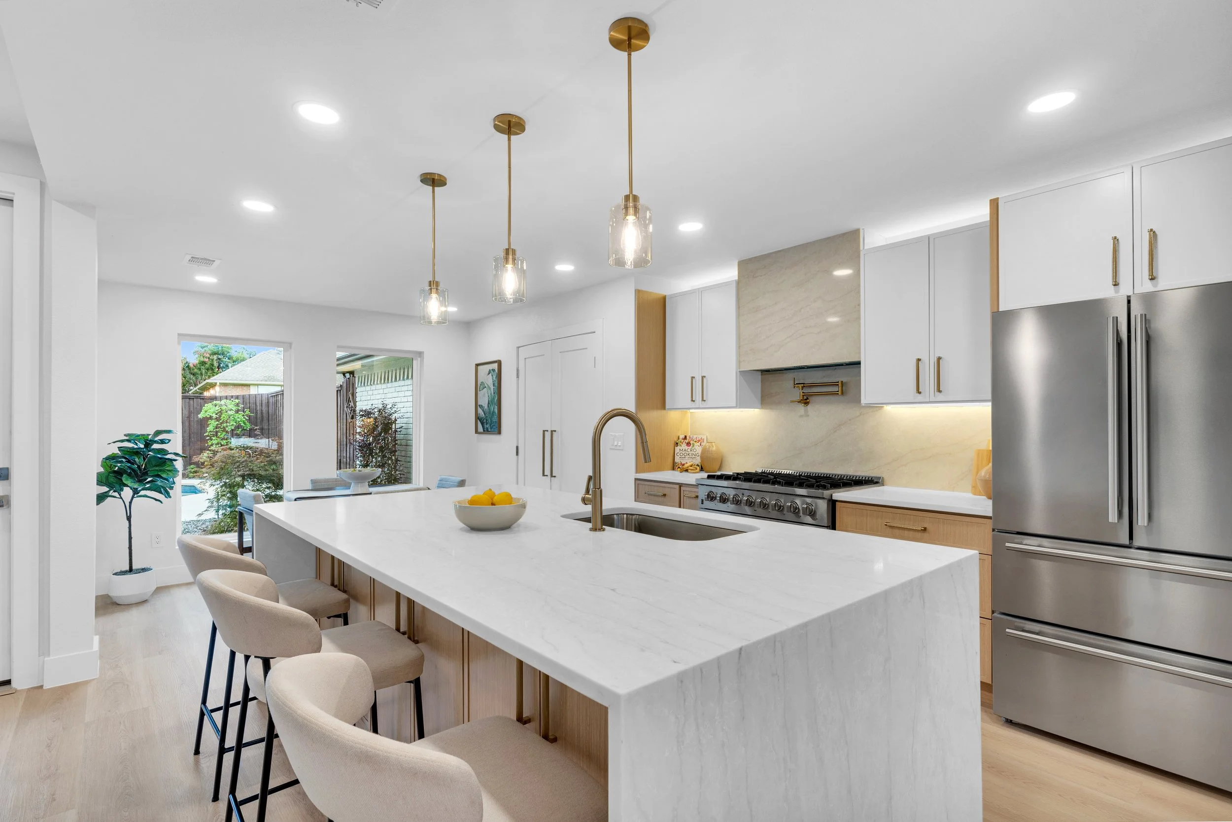 Modern kitchen with an island, beige chairs, pendant lights, white cabinets, stainless steel refrigerator, window with backyard view, and light wood flooring.