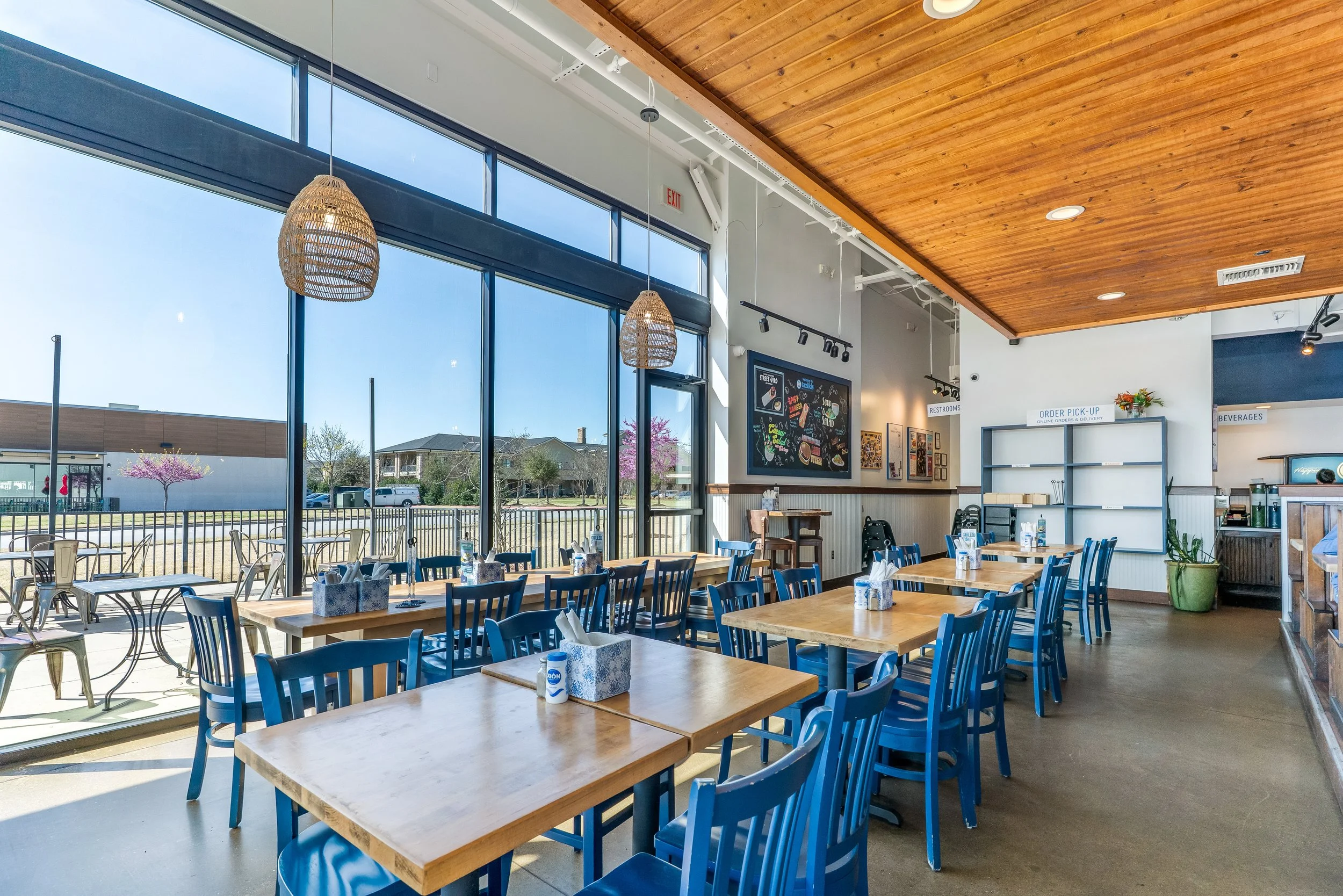 Interior of a bright restaurant with large windows, wooden ceiling, and blue chairs around light wooden tables, set with napkins and salt shakers, with outdoor seating visible through the windows.