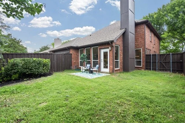 Backyard view of a brick house with a gray roof, patio furniture, and a fenced yard with green grass and trees.