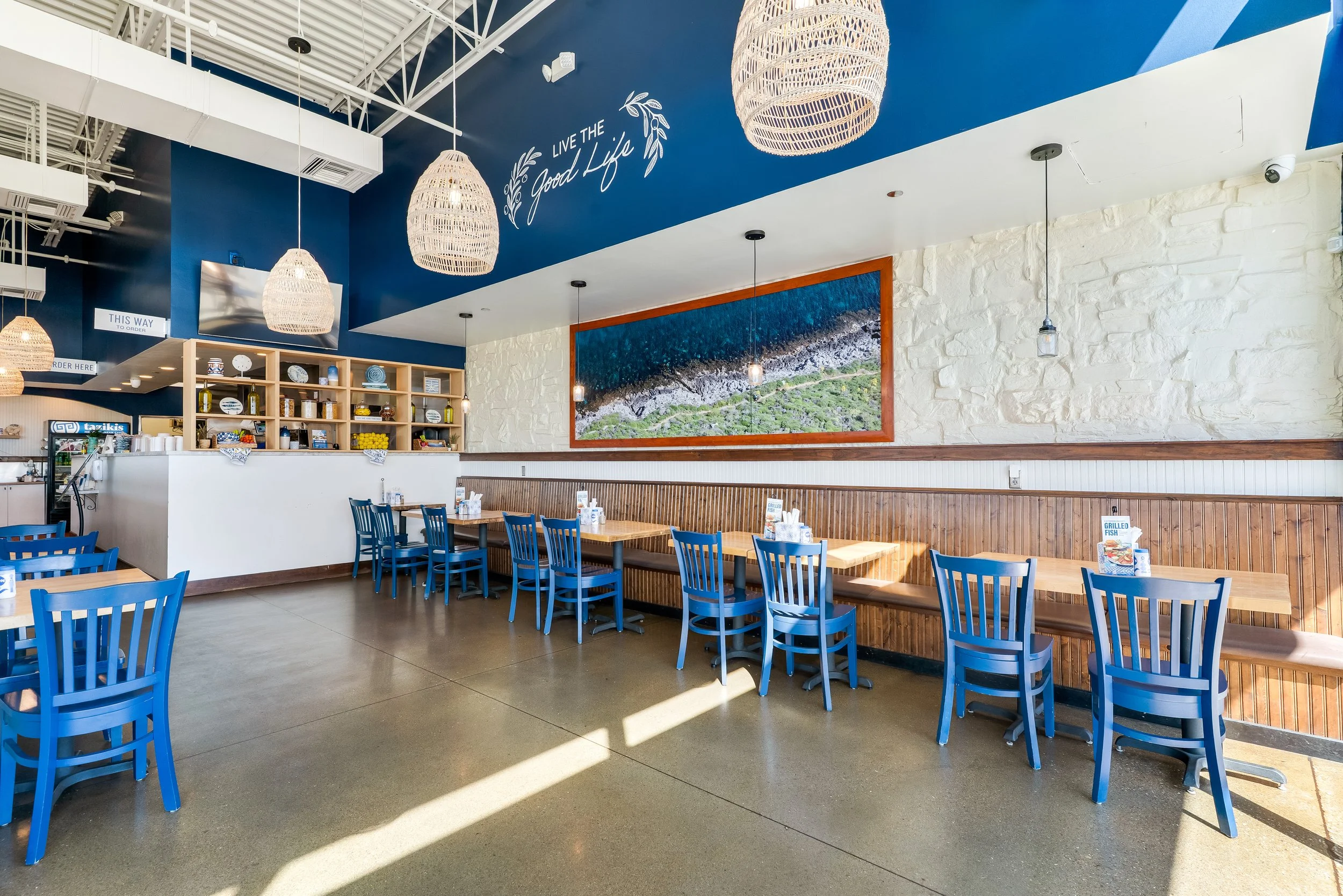 Interior of a restaurant with blue chairs, wooden tables, a white brick wall, and a large landscape photograph on the wall. Woven hanging light fixtures and a sign that says 'Live the Good Life' are visible.