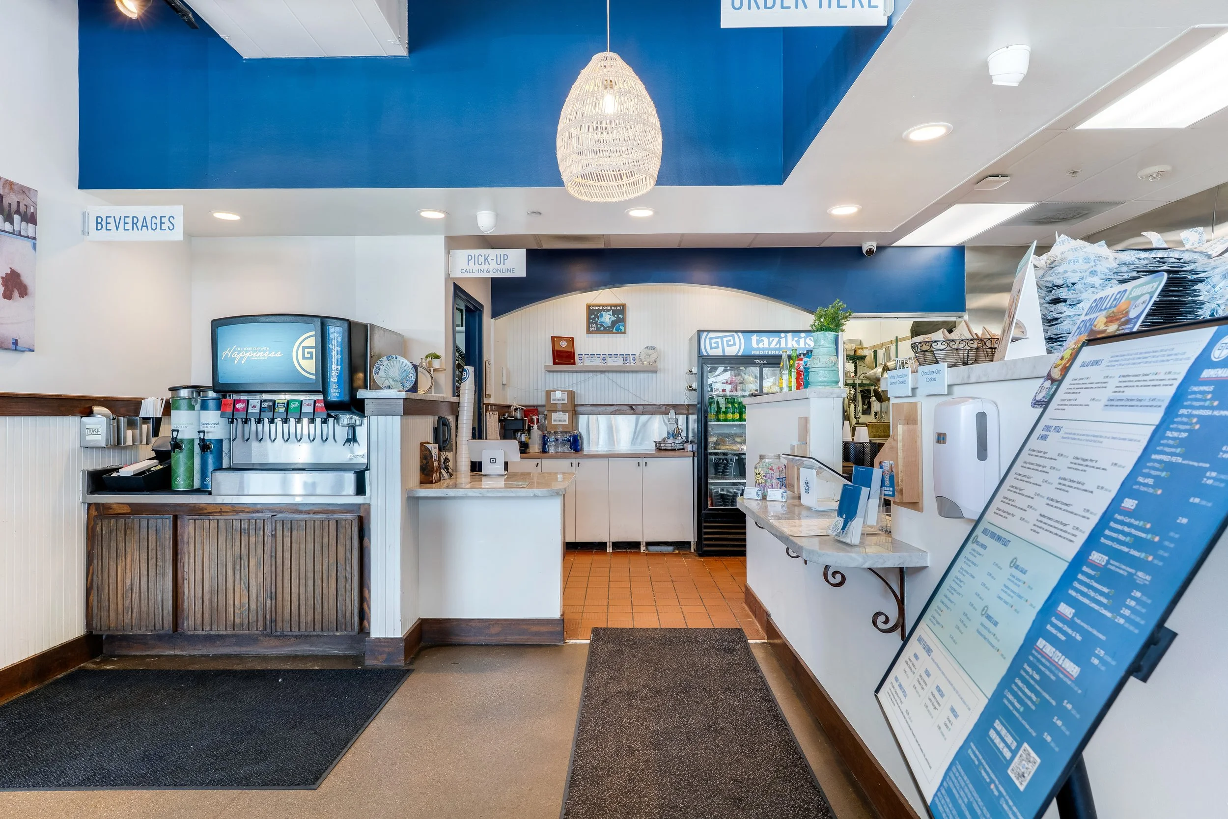 Interior of a restaurant counter with beverage dispensers, menu boards, and a door labeled 'Pick Up'. The decor features a blue and white color scheme with a hanging wicker light fixture and framed artwork on the wall.