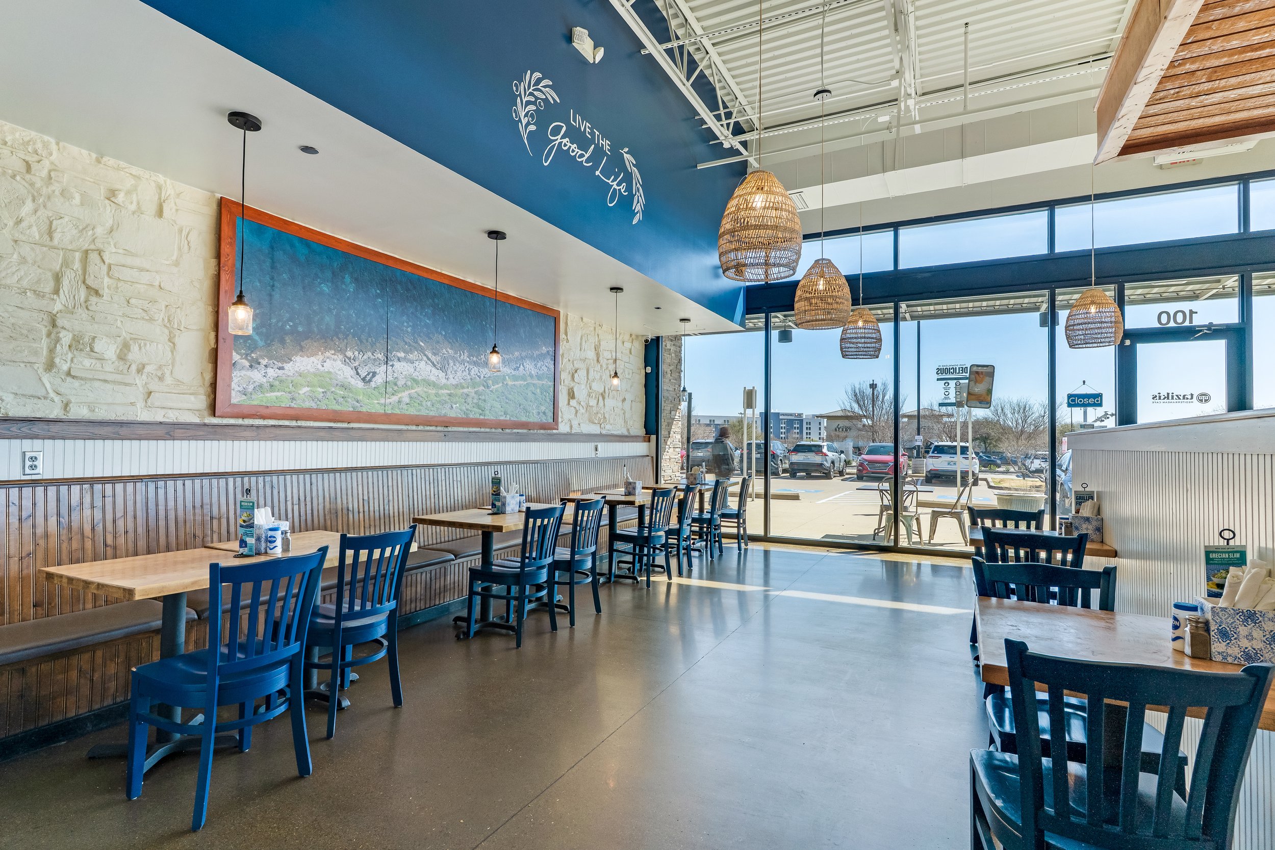 Inside a bright casual restaurant with wooden tables, blue chairs, wicker pendant lights, a large window, and a blue wall with the phrase 'Live the Good Life' and a mountain landscape picture.