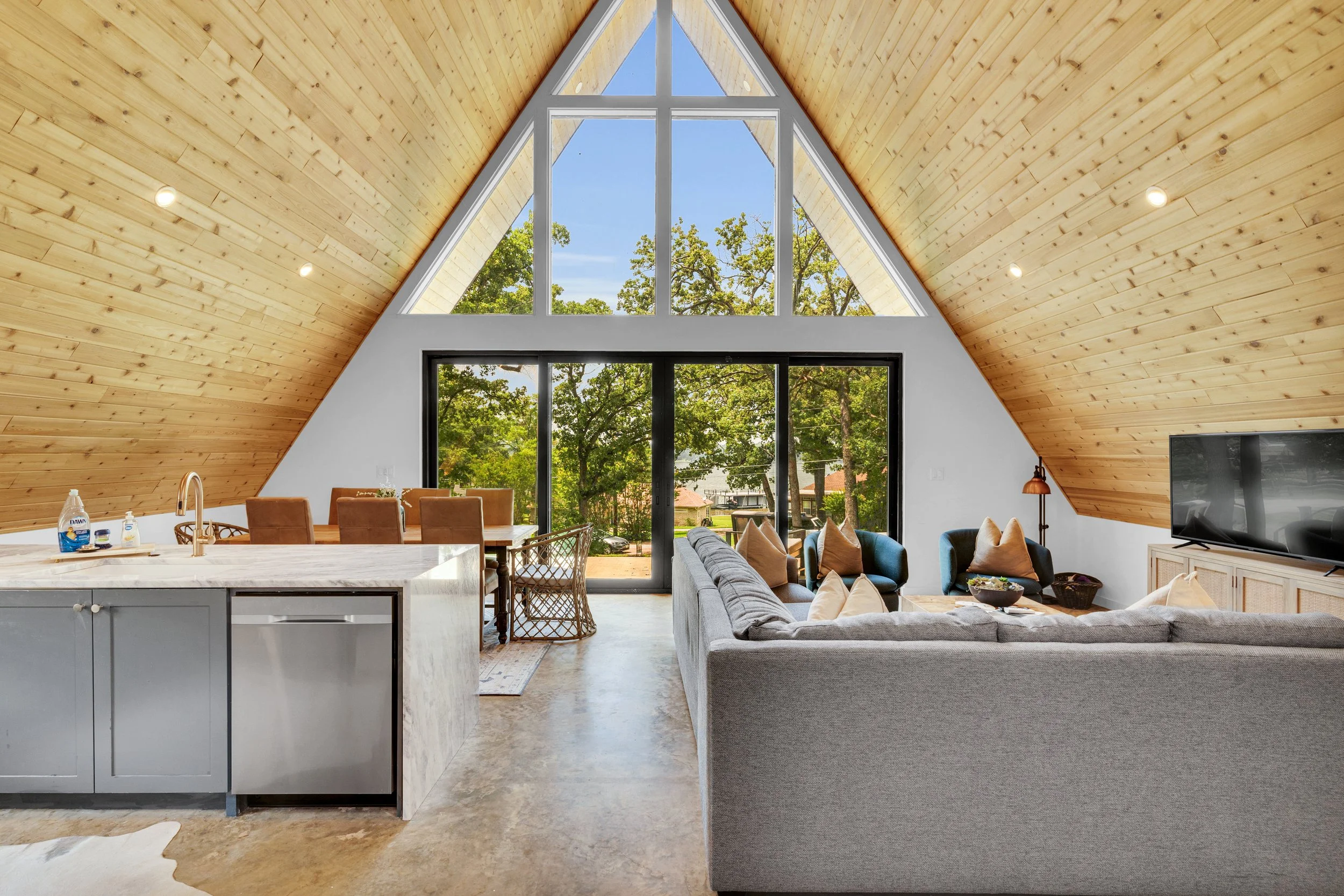 Interior view of a modern living room with a high, triangular wooden ceiling and large windows showing trees outside. The room has a gray sectional sofa with multiple cushions, a flat-screen TV, and a dining area with a marble-topped table and chairs. Bright natural light illuminates the space.