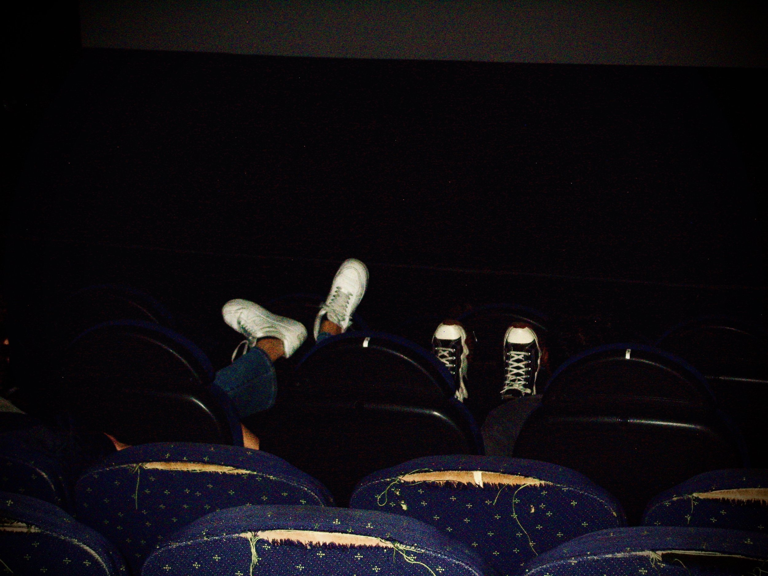 Photo of people's legs and shoes while sitting in dark auditorium, with visible exit sign at the top