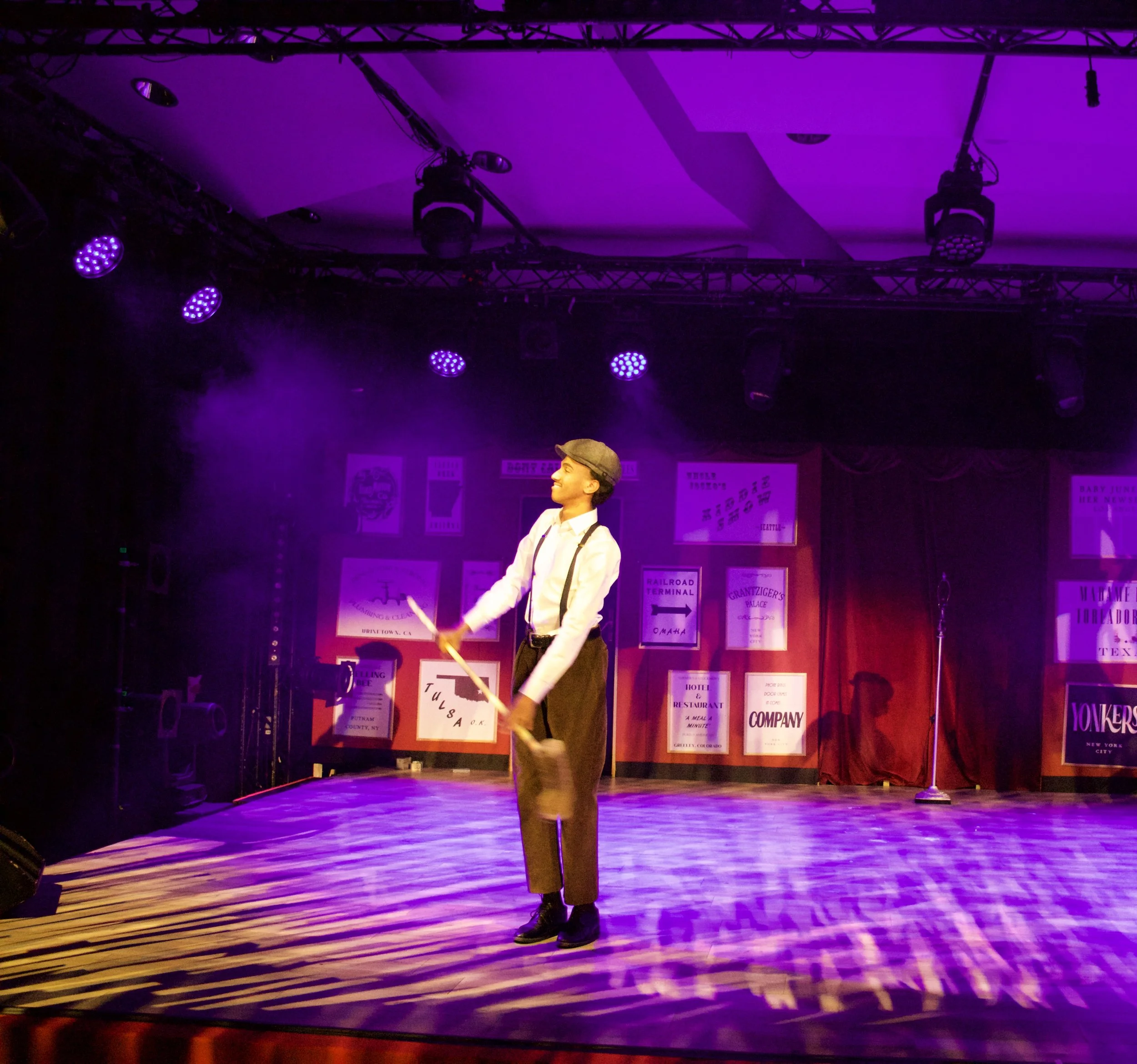 A performer in vintage clothing, including suspenders and a flat cap, is spinning a hula hoop on stage under purple and yellow stage lights.