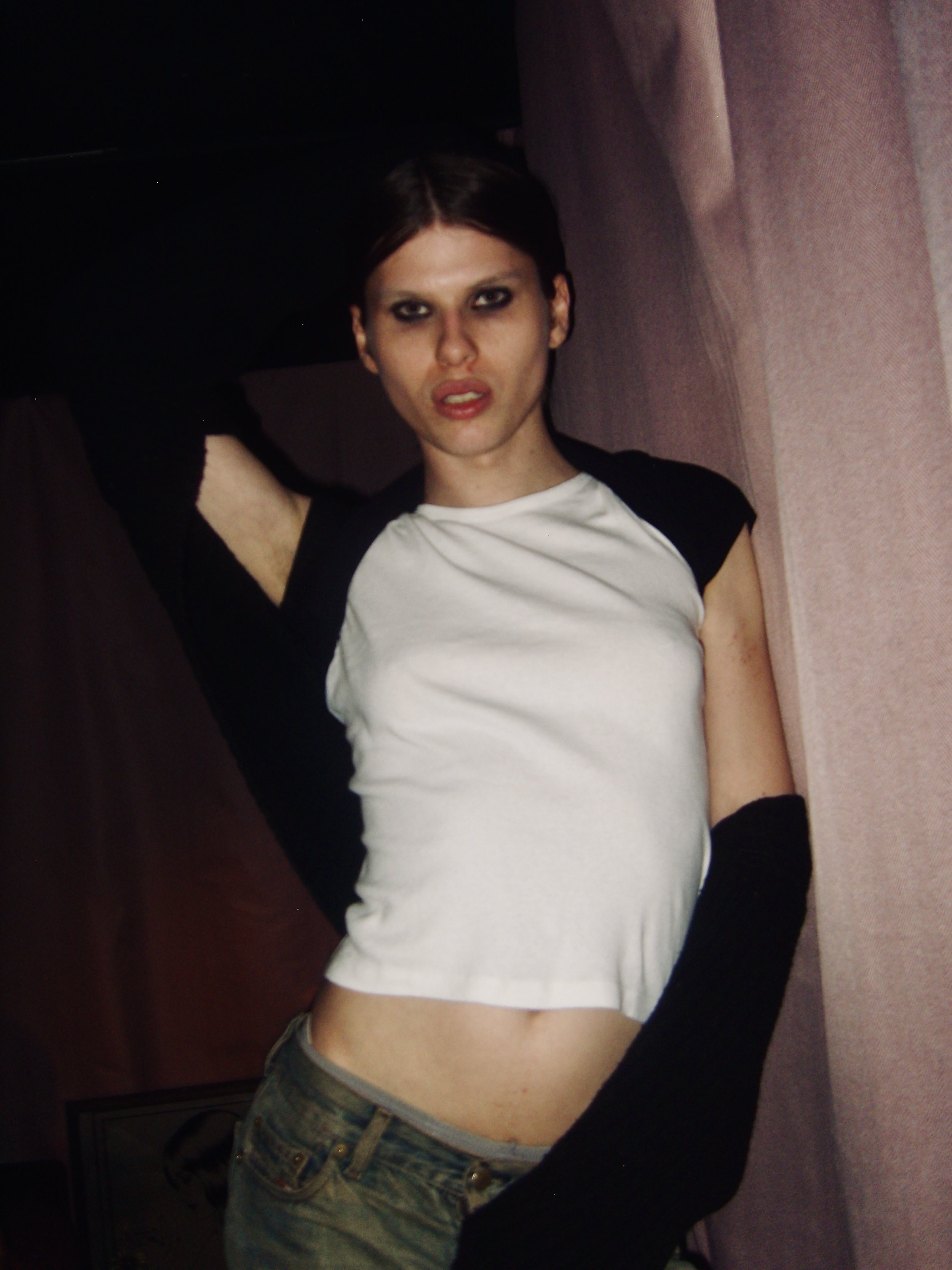 A person with short dark hair and smokey eye makeup wearing a white and black baseball-style shirt, camouflage pants, and a belt, sitting on a pinkish-purple couch with armrest, in a dimly lit room.