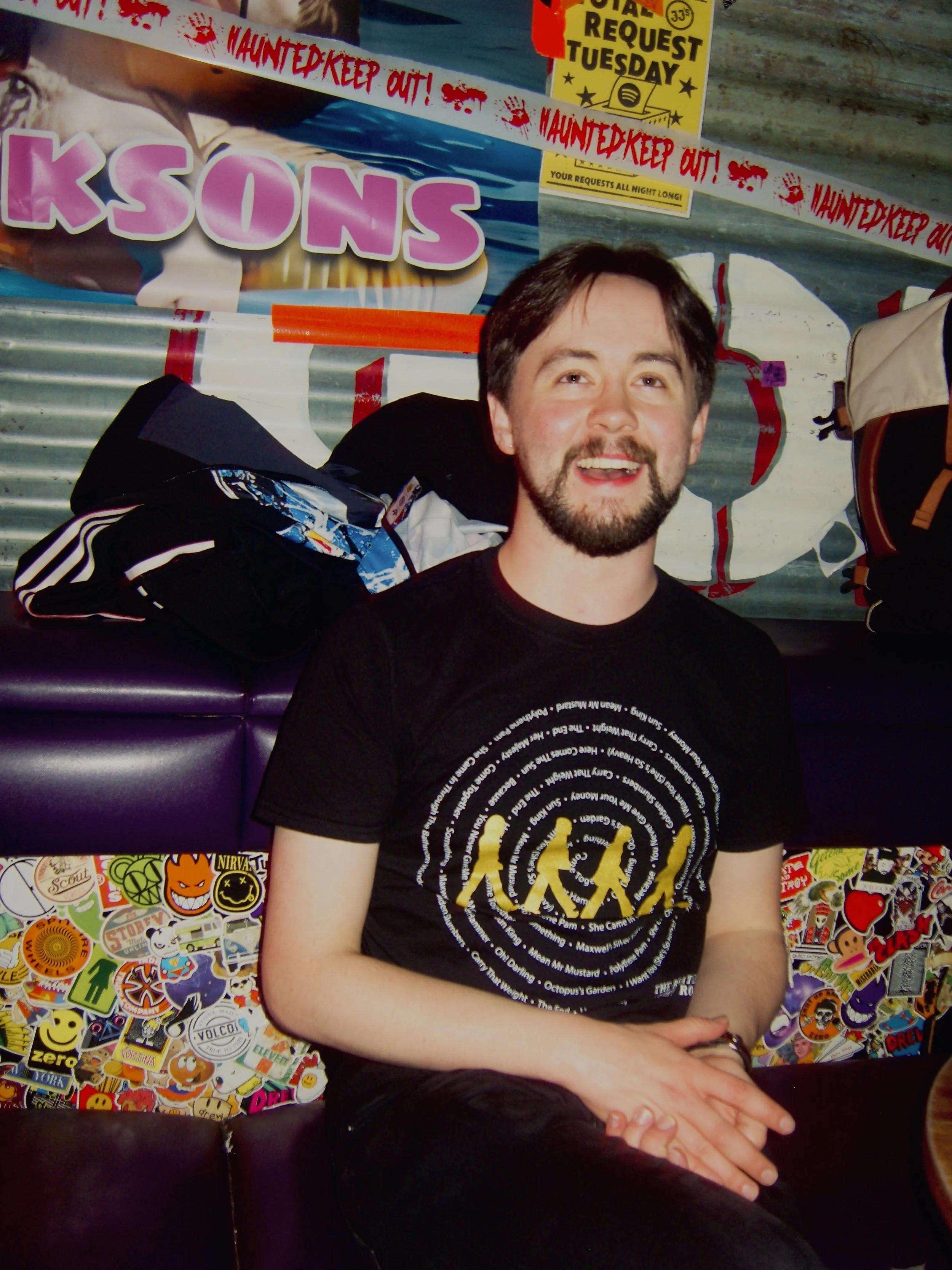 A man with a beard and short dark hair smiling, wearing a black t-shirt with yellow text and a circular design, sitting in front of a wall decorated with colorful stickers and posters.