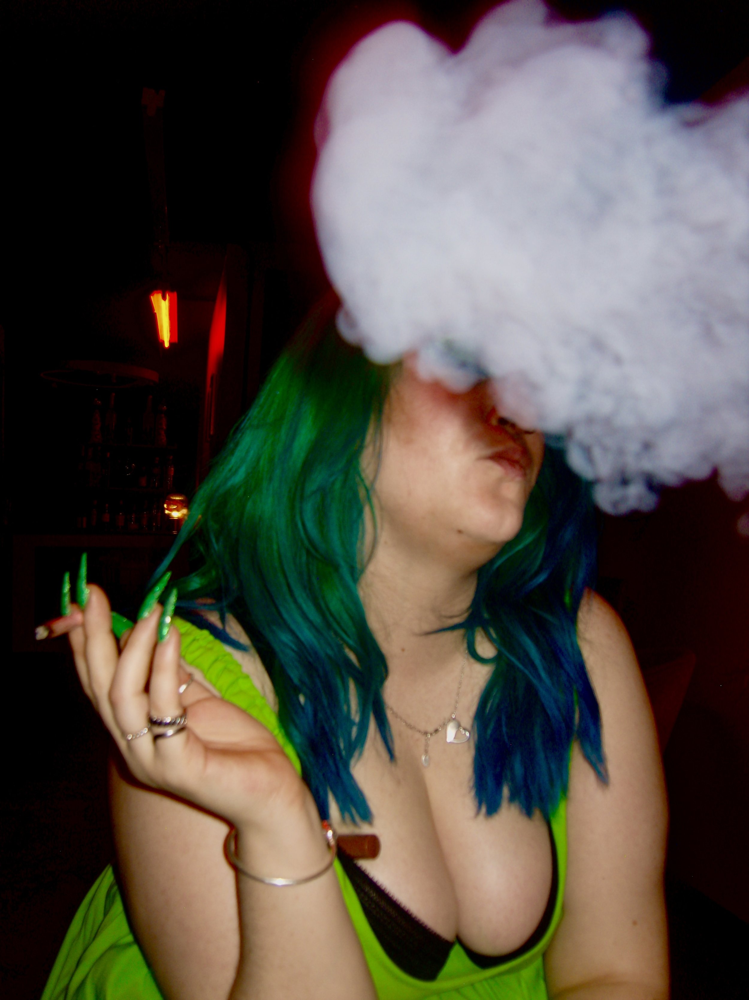 A woman with bright green and blue hair, wearing a neon green dress and multiple rings, with smoke partially obscuring her face, in a dimly lit room with red and green lighting.