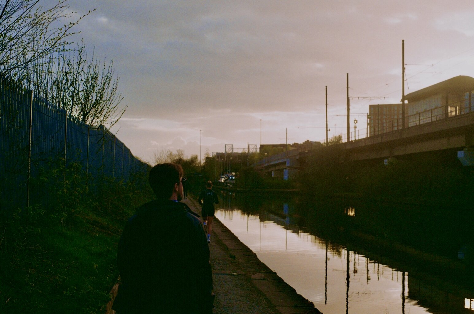 People jogging along a canal under a cloudy sky, with a bridge in the distance and a fence on the left side