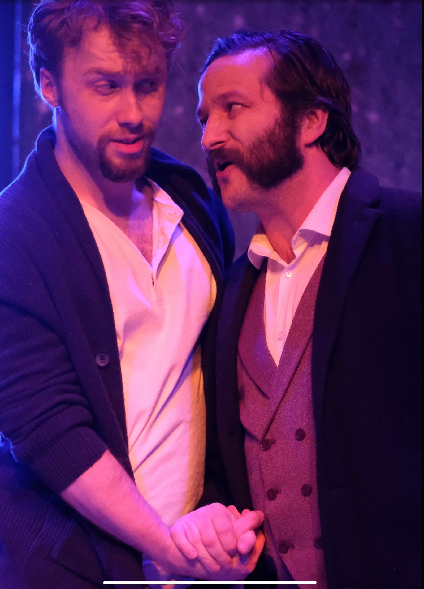 Two men shaking hands and smiling, dressed in suits and shirts, with purple lighting in the background.