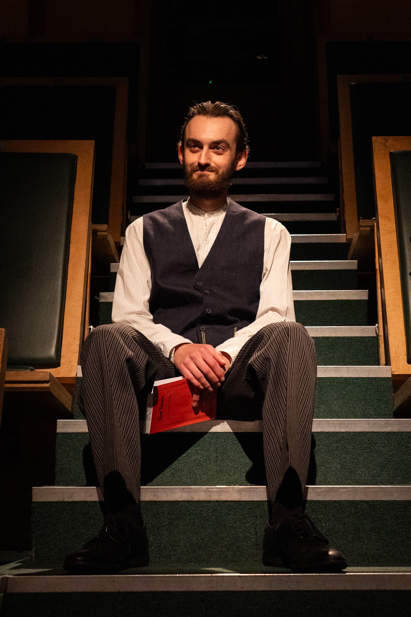 A man with a beard and slicked-back hair sitting on stairs in a dark room, holding a red packet, wearing a vest over a long-sleeved shirt with striped pants, looking slightly smiling.