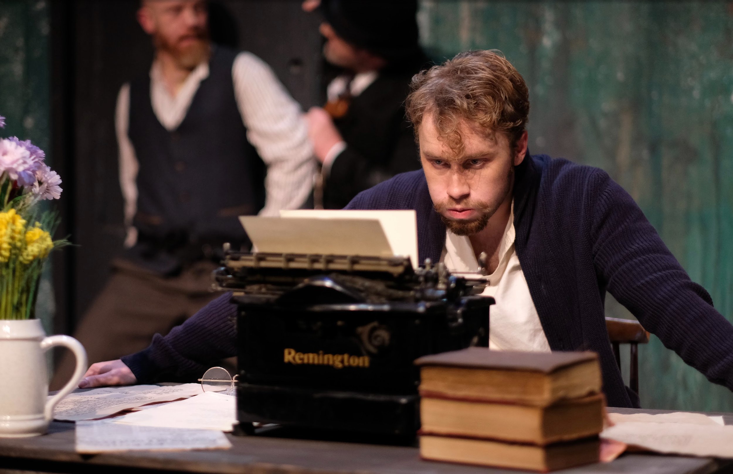 A young man with curly hair and a beard sitting at a table, focused on a typewriter in front of him. There are books stacked on the right side of the table and flowers on the left. In the background, two men are engaged in a conversation, out of focu