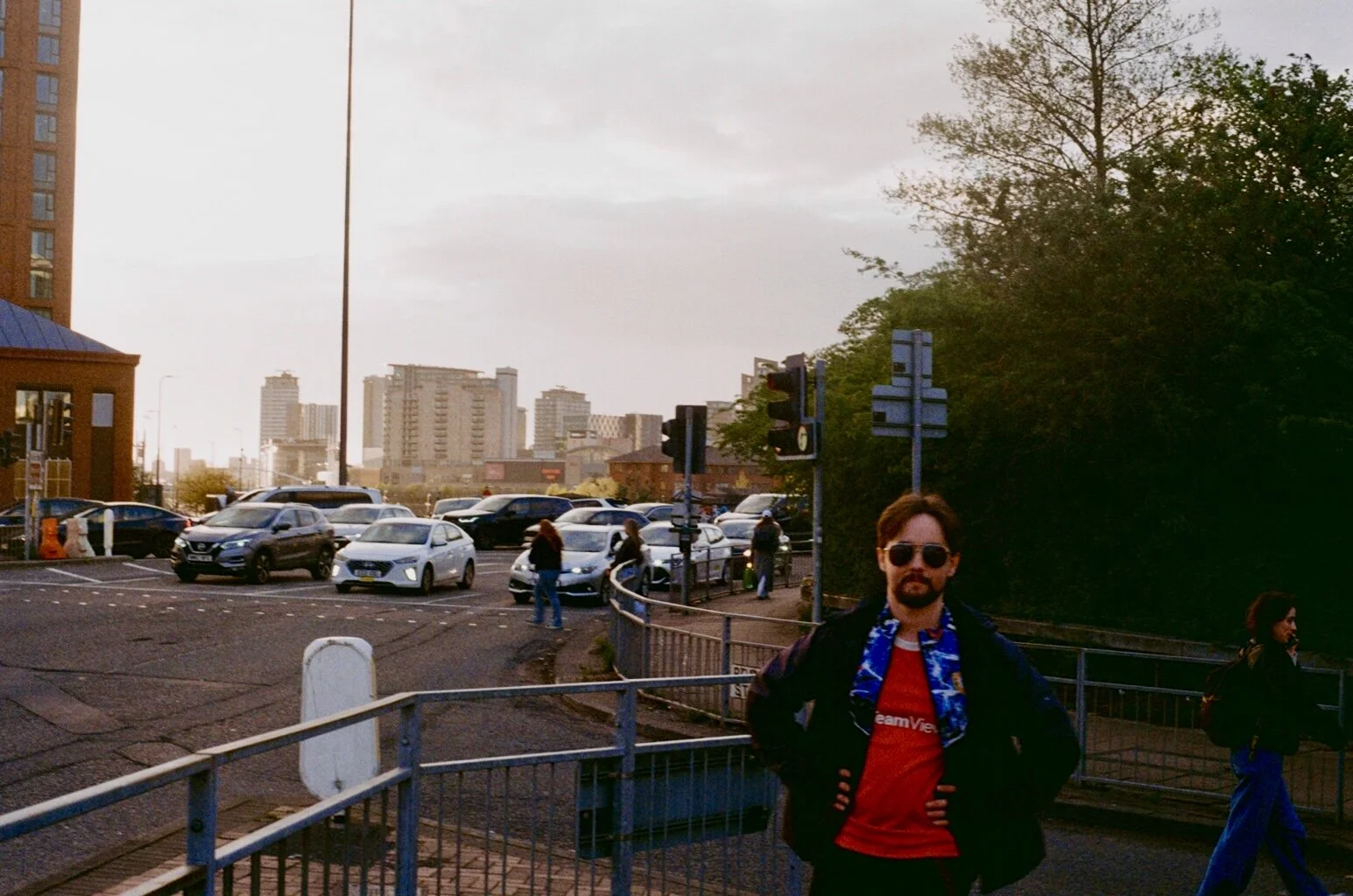A man with sunglasses, a beard, and dark hair stands with his hands on his hips in front of a parking lot with cars and a cityscape in the background. The scene is during sunset or early evening.