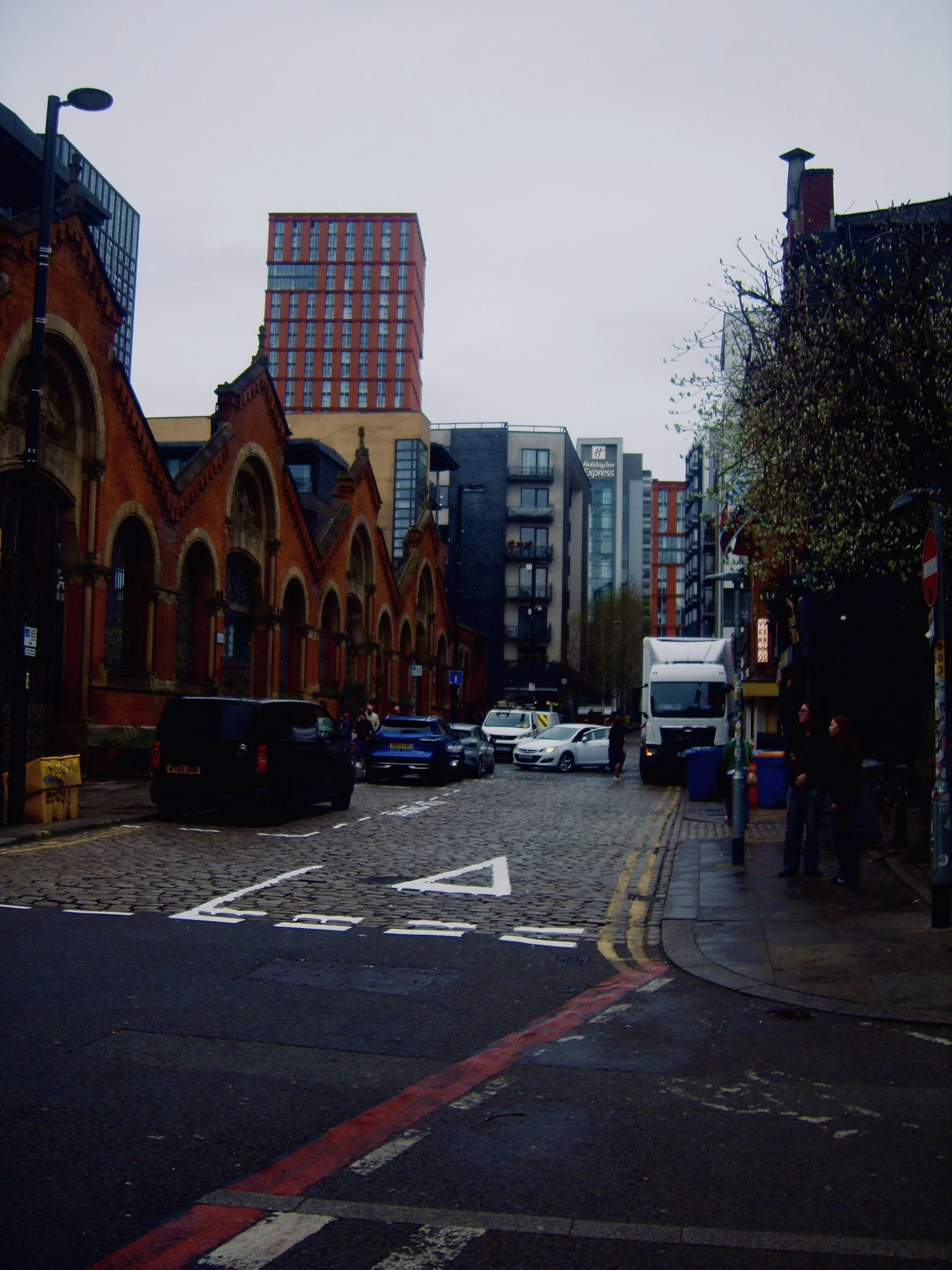 A city street with cars parked along the cobblestone road, historic red-brick buildings with arched windows on the left, modern high-rise buildings in the background, street pedestrians, and trees on the right.