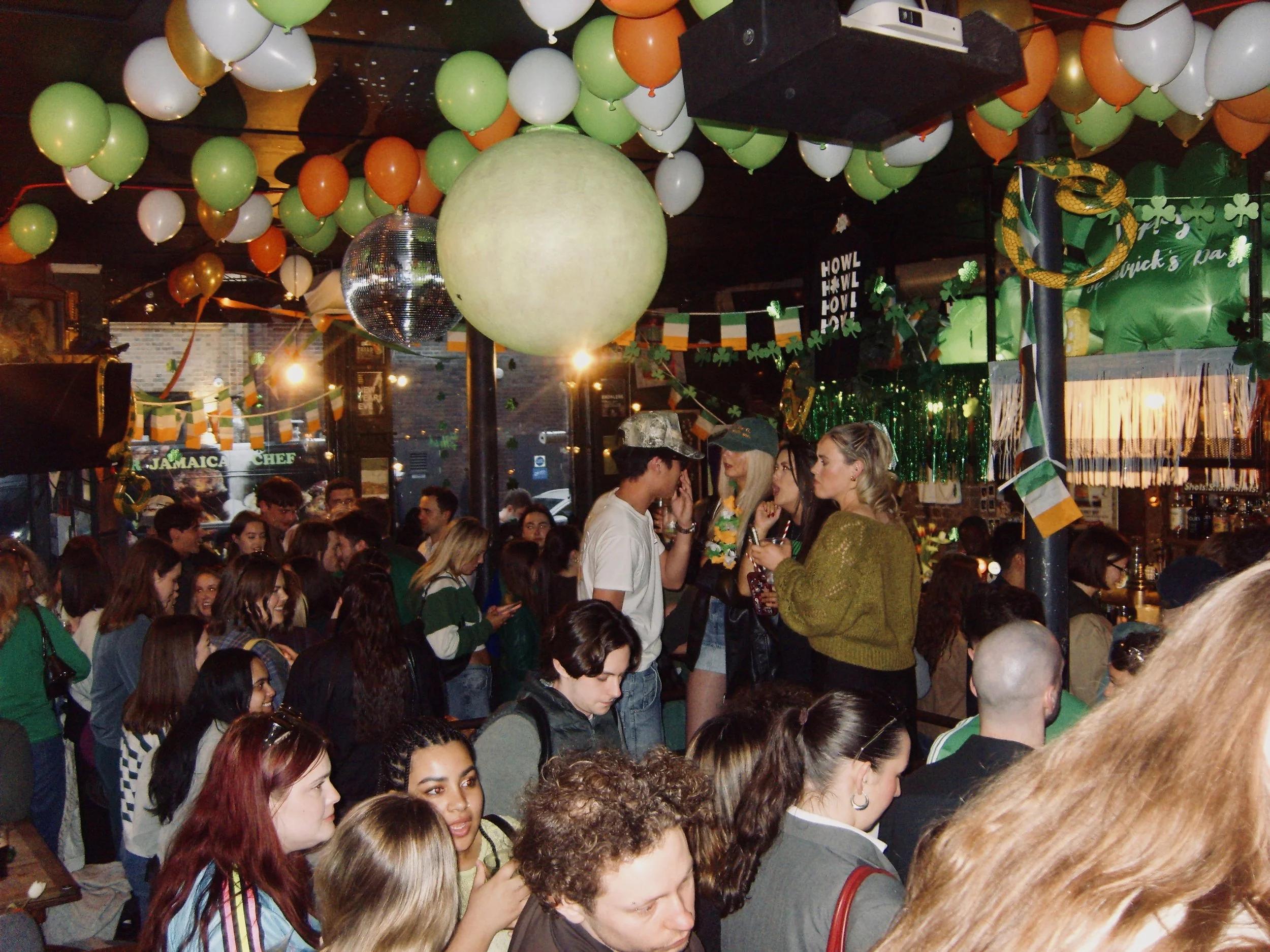 Crowd of people celebrating St. Patrick's Day with green, white, orange balloons and decorations, including shamrocks, in a festive indoor setting.