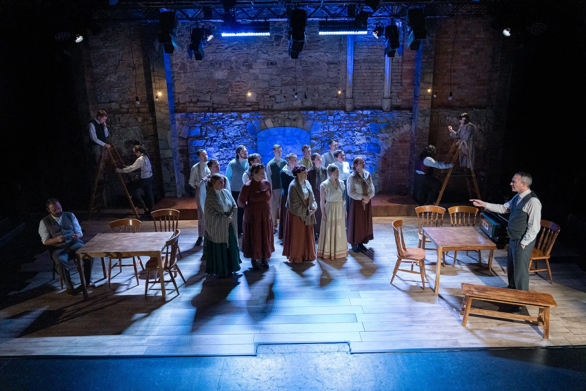 A group of performers on a stage rehearsing a scene, with some seated and others standing, under blue stage lighting in front of a stone wall, with a conductor or director in the foreground.