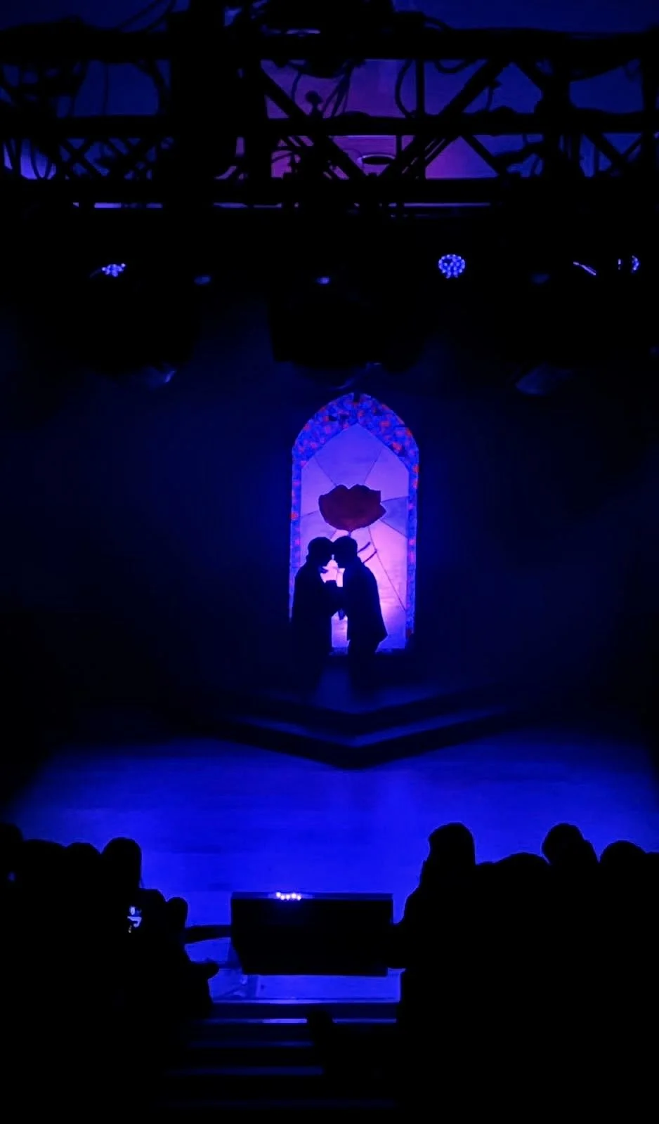 Silhouettes of two people facing each other holding hands in front of a colorful stained glass window with a large red flower, illuminated in purple and blue lighting, on a stage with an audience in the foreground.
