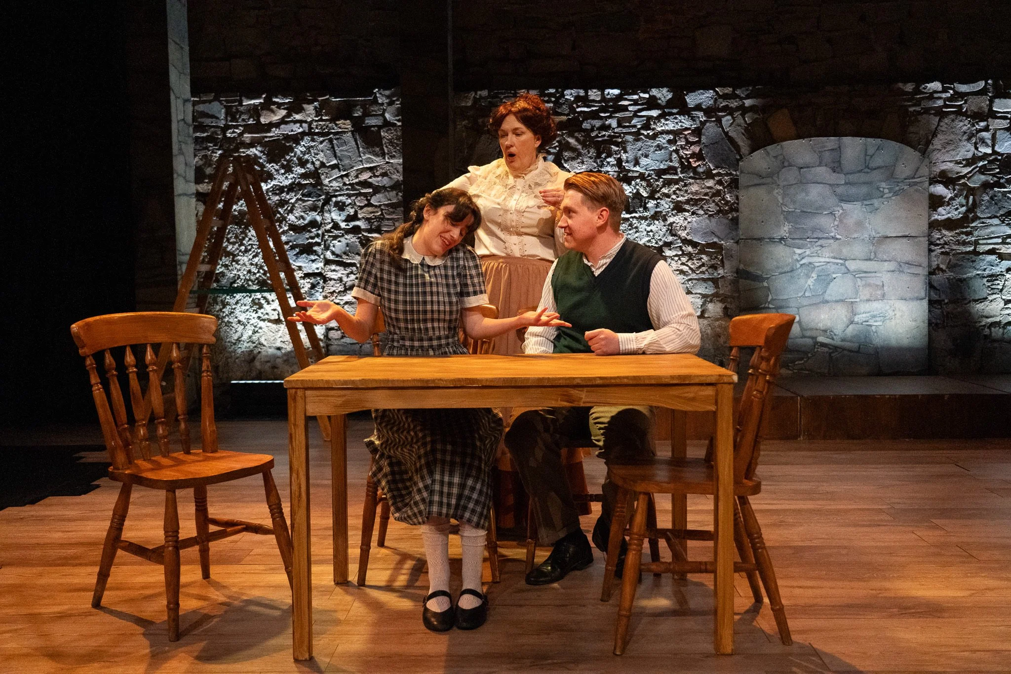 Three women on stage performing a scene. One woman stands behind two women sitting at a wooden table, appearing to give instructions or conduct. The women are dressed in vintage clothing, and the setting resembles an old-fashioned kitchen or living r